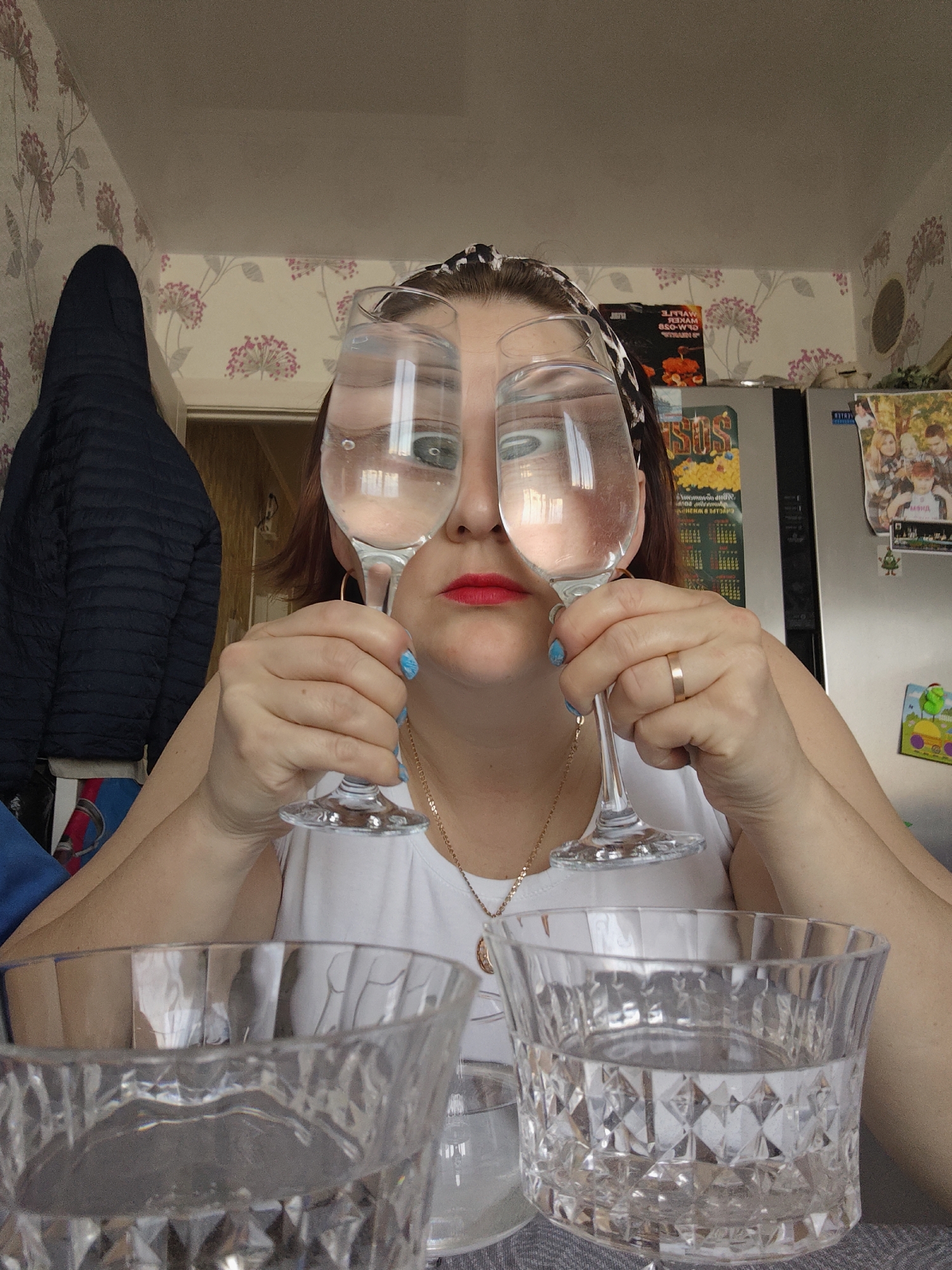 The image captures a playful indoor scene, likely in a home kitchen or dining area in Markovskii, Russia. The main subject is a woman, viewed from a mid-shot perspective, holding two clear wine glasses to her eyes. The glasses, which appear to contain clear liquid, distort and enlarge her eyes, suggesting a humorous or whimsical pose as she looks directly at the camera. She has red lipstick, blue nail polish on her left hand, and a simple gold band ring on her right hand. She is wearing a white top, a patterned headband, and a gold chain necklace.
In the foreground, on a grey textured surface, are two large, faceted glass bowls or tumblers, both filled with clear liquid.
The background features domestic details. To the left, a dark blue quilted jacket hangs on a hook against wallpaper decorated with a repeating floral pattern of pink blossoms and green leaves on a light background. To the right, a silver refrigerator is visible, covered with an assortment of magnets, several personal photographs, and a colorful calendar. Text visible on the calendar includes Russian words such as "ЯНВАРЬ" (January), "МАРТ" (March), and "СУББОТА" (Saturday). Above the refrigerator, a black package or box displays partially legible Russian text, including "ПЕКАРЬ" (Baker) and "ГЛЮКОЗА" (Glucose). The lighting is bright and even, indicating daytime conditions.