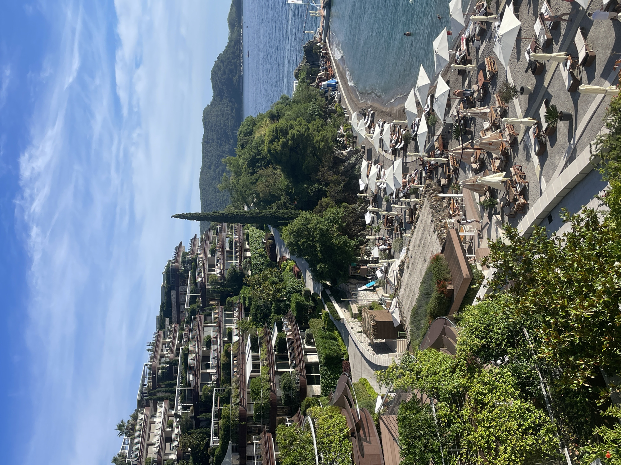 A panoramic, high-angle shot captures a luxury resort complex built into a hillside overlooking a pebble beach and the Adriatic Sea in Budva, Montenegro. The resort's architecture features multiple terraced levels, with each level adorned with lush greenery, private balconies, and white awnings. The beach area below is furnished with numerous white lounge chairs and umbrellas, creating a vibrant, sun-drenched scene. Several people are visible on the beach, relaxing under the umbrellas or swimming in the clear blue water. A pier extends into the sea, with a few boats moored nearby. The surrounding landscape is dominated by rolling green hills, a clear blue sky with scattered white clouds, and the tranquil expanse of the sea. The overall impression is one of serene, upscale coastal relaxation.