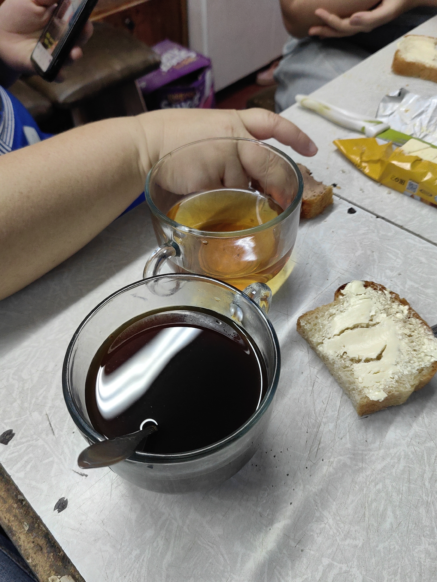 A group of people are having a snack at a table in Shymkent, Kazakhstan. In the foreground, two glass cups are filled with dark liquid, possibly coffee or tea. A slice of bread with butter is on the right, next to a foil package of butter. In the background, someone is holding a smartphone, and a purple bag with an unreadable label is visible. The table appears to be made of a light-colored, textured material, and there are some crumbs and smudges on its surface. The lighting suggests an indoor setting, possibly a home or a cafe.