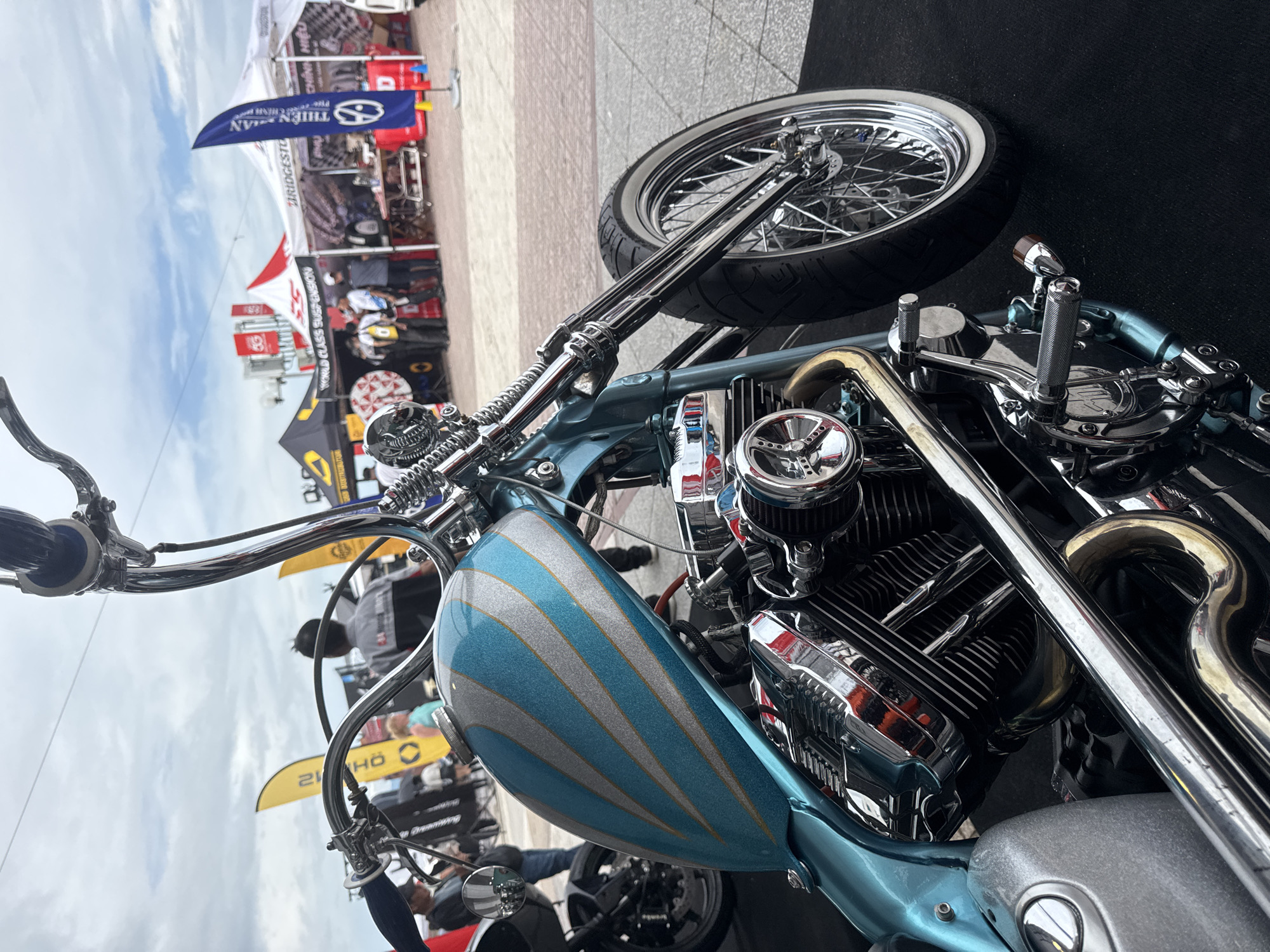 The image shows a detailed view of a custom motorcycle, likely showcased at an event or gathering. The motorcycle features a distinctive light blue gas tank with silver stripes and lots of polished chrome elements. Parts of the engine are visible, as are exhaust pipes, handlebars, and the front wheel with its spokes. In the background, there are tents and people suggesting a public event or exhibition. There's no indication of the city name, therefore location is unknown.