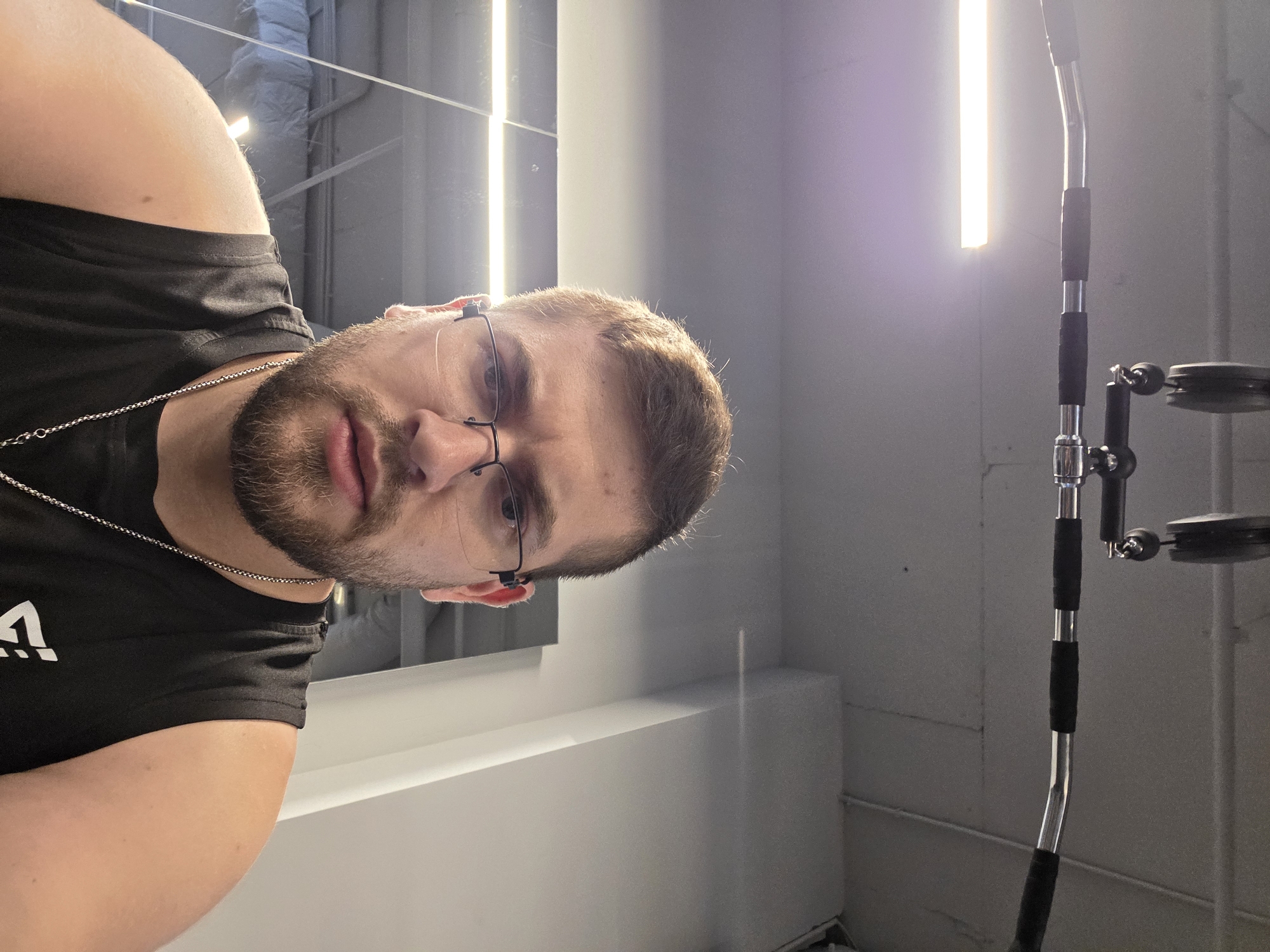 A man with short brown hair, a beard, and glasses is looking directly at the camera. He is wearing a black t-shirt with a white logo. A silver chain is visible around his neck. He appears to be in a gym, as indicated by the gym equipment in the background. The background includes a light fixture and what appears to be a cable machine. The location cannot be determined from the provided image; there is no evidence to suggest this image was taken in Plovdiv, Bulgaria.