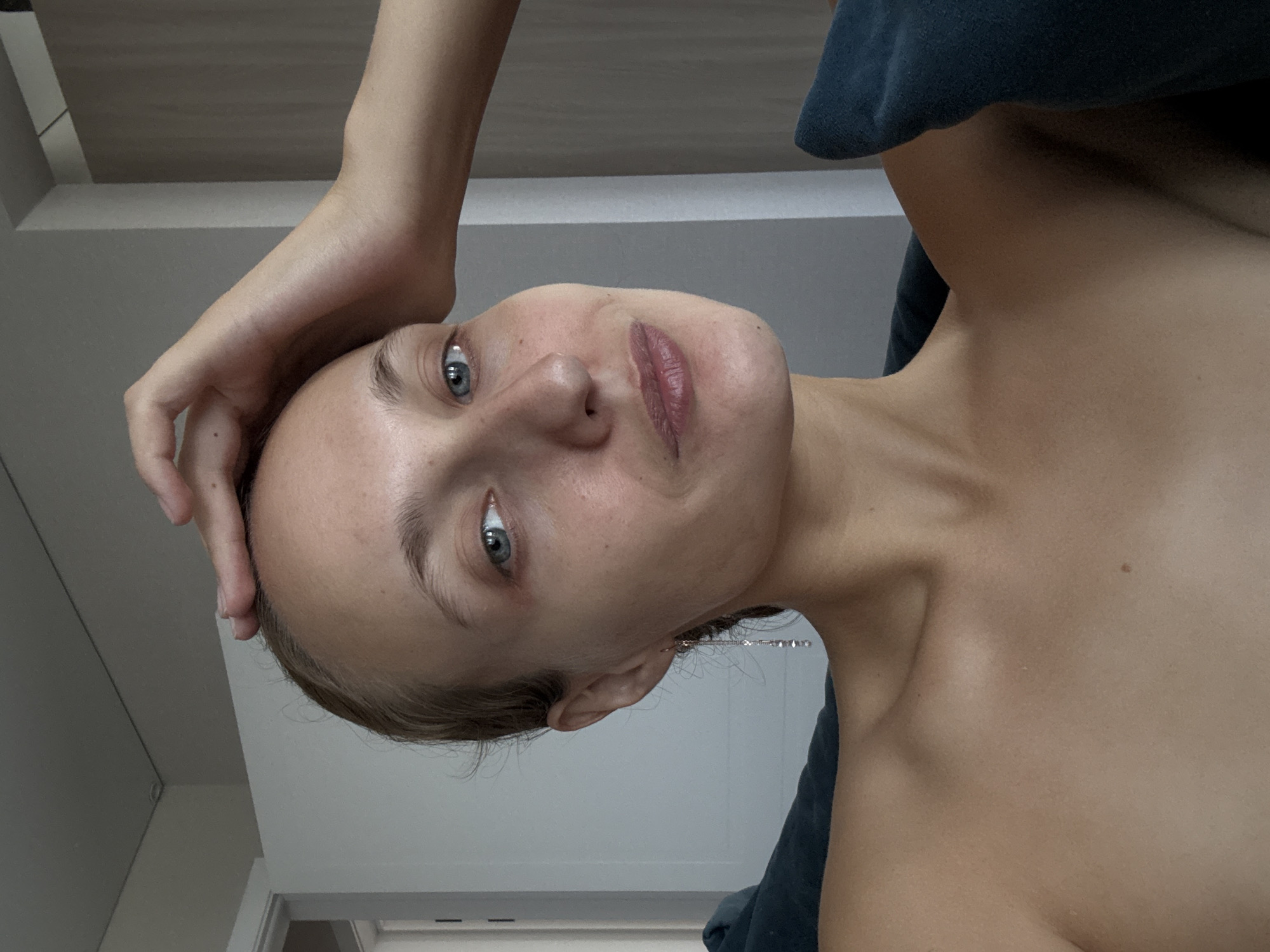 A young woman is pictured from the chest up, indoors. Her left arm is raised, with her hand resting on the top of her head, fingers spread. Her head is tilted slightly to her right. She has blue eyes and fair skin with a few freckles visible on her cheeks. A long, delicate earring with small clear stones hangs from her left ear. She is wearing a dark blue, plush fabric garment. The background appears to be a room with white walls and a closed door visible on the left. The lighting suggests it is daytime. There is no indication of the specific location within the city of Podshivalovo, Russia.