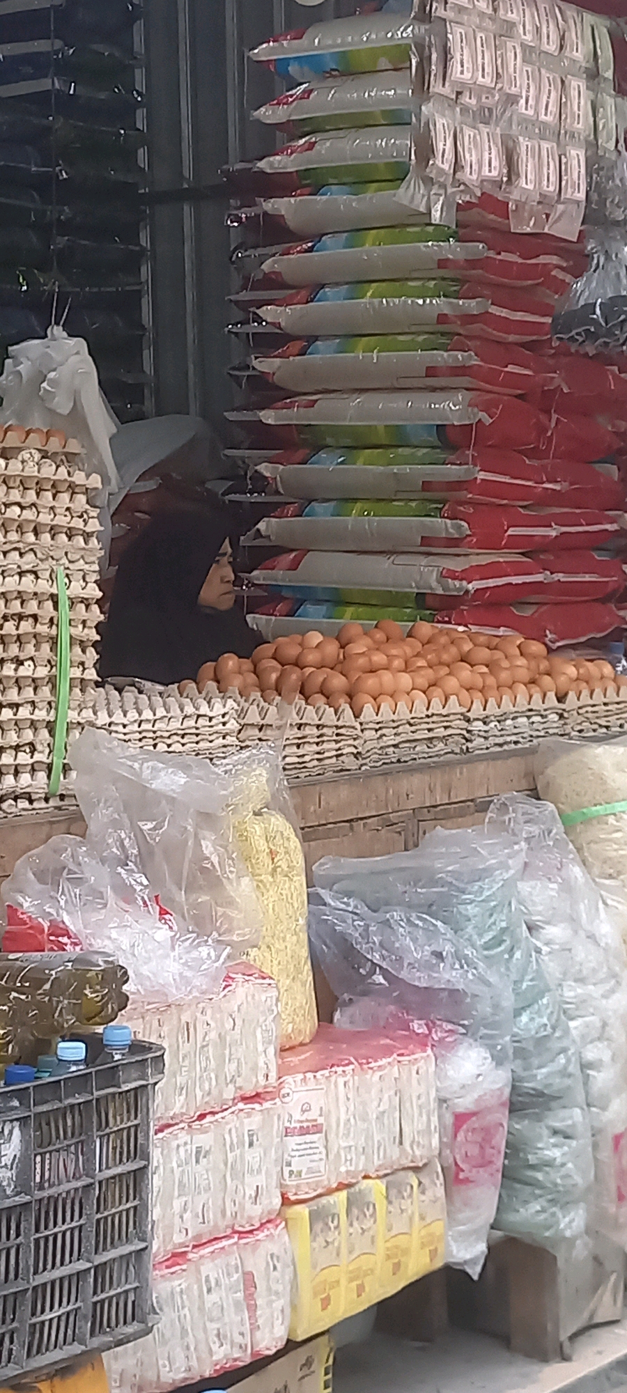 The image captures a street market scene in Melayu, Indonesia, likely a stall selling various food items. In the foreground, a large pile of brown eggs is displayed in cardboard cartons. To the left, stacks of egg cartons are visible, with a person wearing a black hijab partially visible behind them. Behind the eggs, there are stacks of bagged goods, appearing to be rice or similar dry food items, with red, white, and green packaging. Some of these bags have printed labels. In the lower part of the image, there are plastic-wrapped packages of what could be noodles or other dry goods, as well as some bottled items in a dark crate on the left. The overall impression is one of a busy marketplace with a focus on essential food commodities.