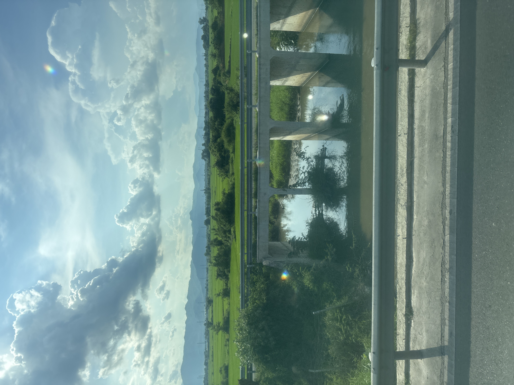 The image captures a scenic view of a rural landscape with a concrete bridge spanning a body of water. Lush green rice fields stretch across the horizon, bordered by distant, hazy mountains and dotted with sparse trees and utility poles. The sky is partly cloudy, with dramatic, sunlit cumulonimbus formations casting shadows on the landscape below. The foreground shows a roadside barrier and a portion of a paved road, suggesting the vantage point is from a vehicle. The water beneath the bridge reflects the sky and the bridge's structure, with some debris visible in the water. Lens flares are present in the upper right and central portions of the image, indicating the presence of a light source.