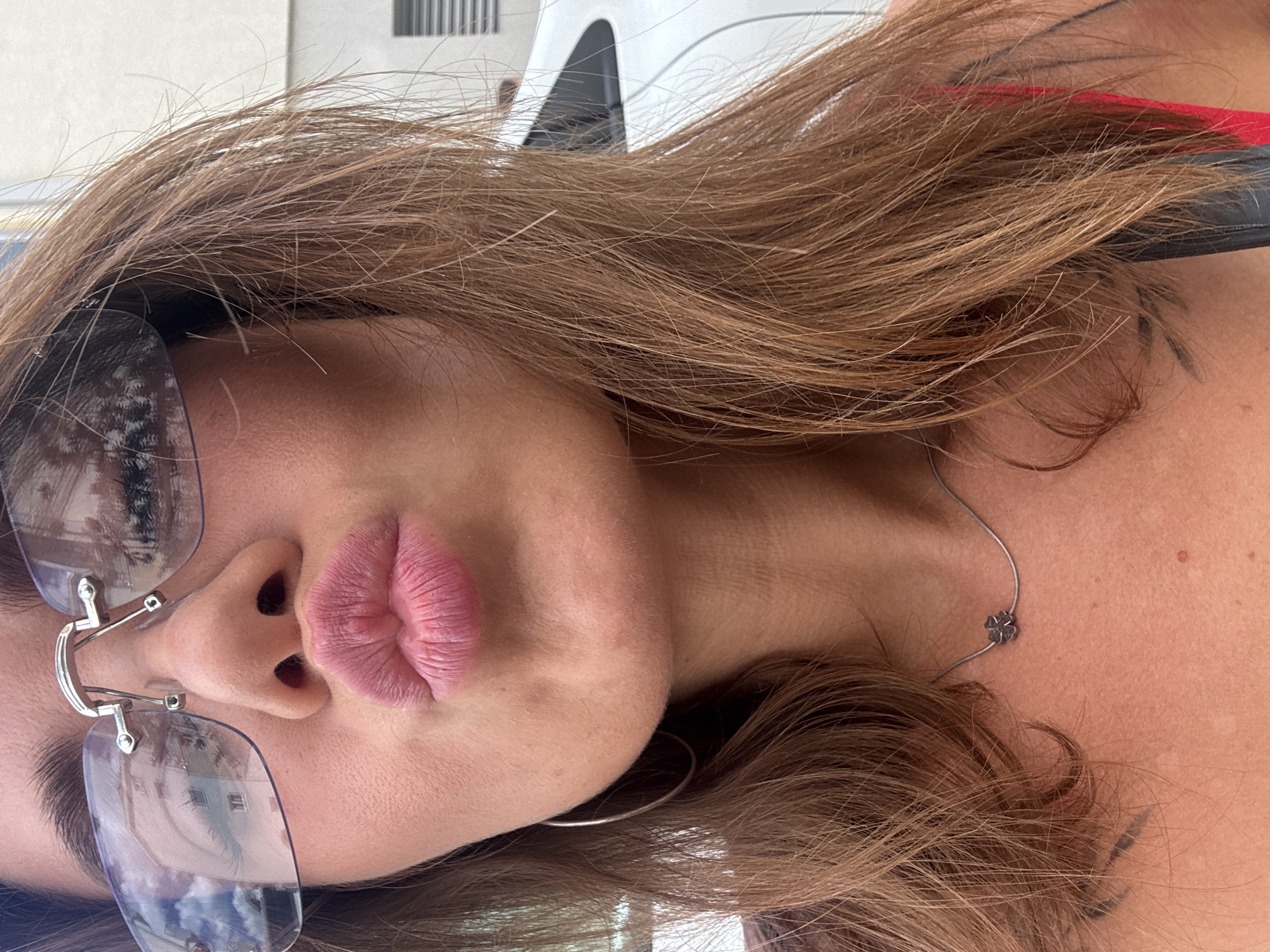A close-up, head-and-shoulders shot of a woman with light brown hair, wearing rimless sunglasses, pouting her lips as if for a kiss. Her hair is parted to the side and falls around her face. She is wearing a necklace with a small, four-leaf clover pendant and large hoop earrings. Reflections of buildings and trees are visible in her sunglasses. The background shows a white car. The overall scene suggests an outdoor setting in Budva, Montenegro.