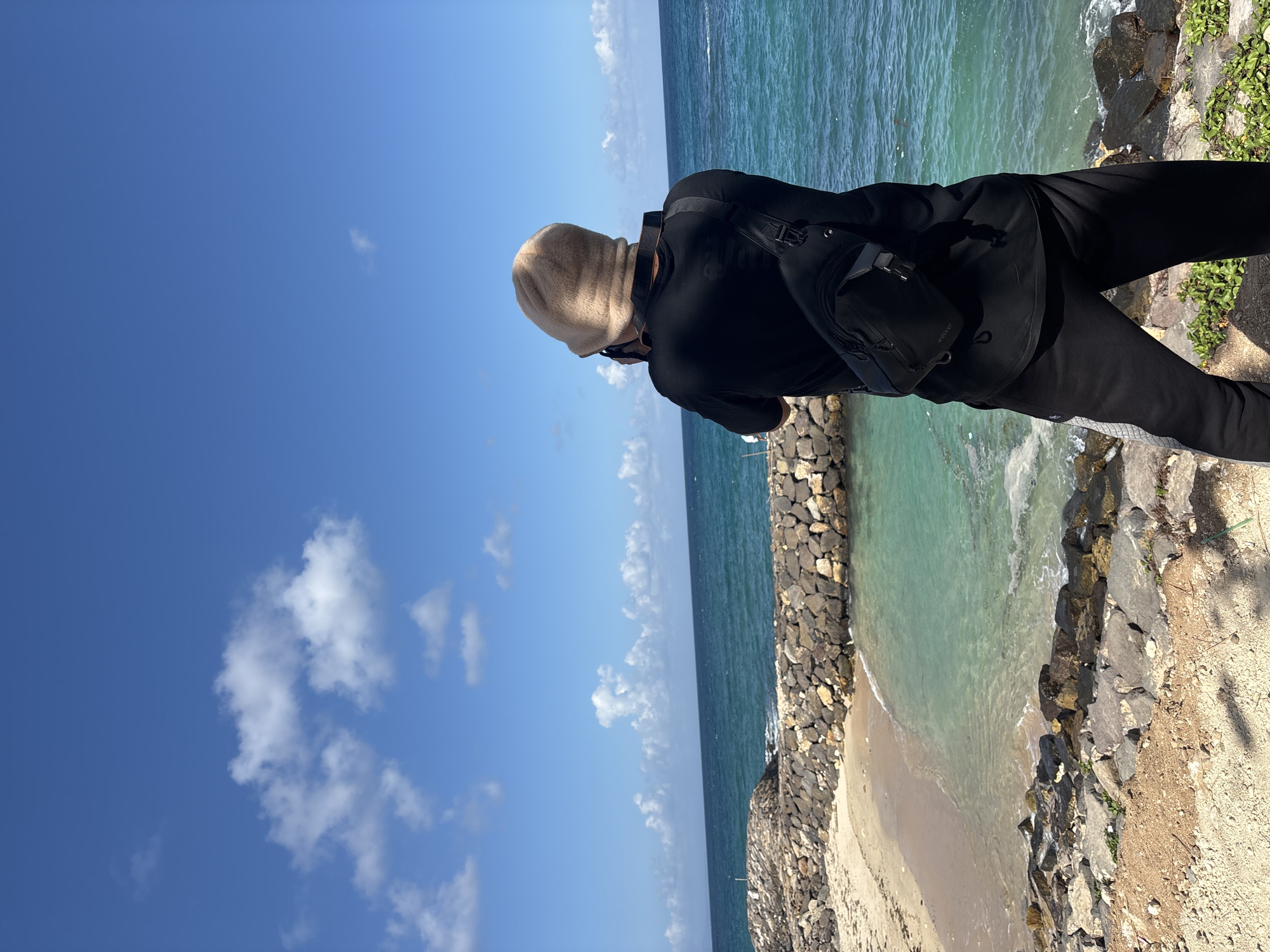 A person stands with their back to the camera, wearing a black t-shirt, black sweatpants, and a beige beanie. They are also wearing a black sling bag across their chest. The person is positioned on a rocky shore, facing a body of turquoise water. In the background, there is a sandy beach bordered by a rock seawall. The sky is blue with scattered white clouds. The scene appears to be a coastal area, possibly the city of Kuta Selatan, Indonesia, given the clear water, sandy beach, and rocky shoreline.