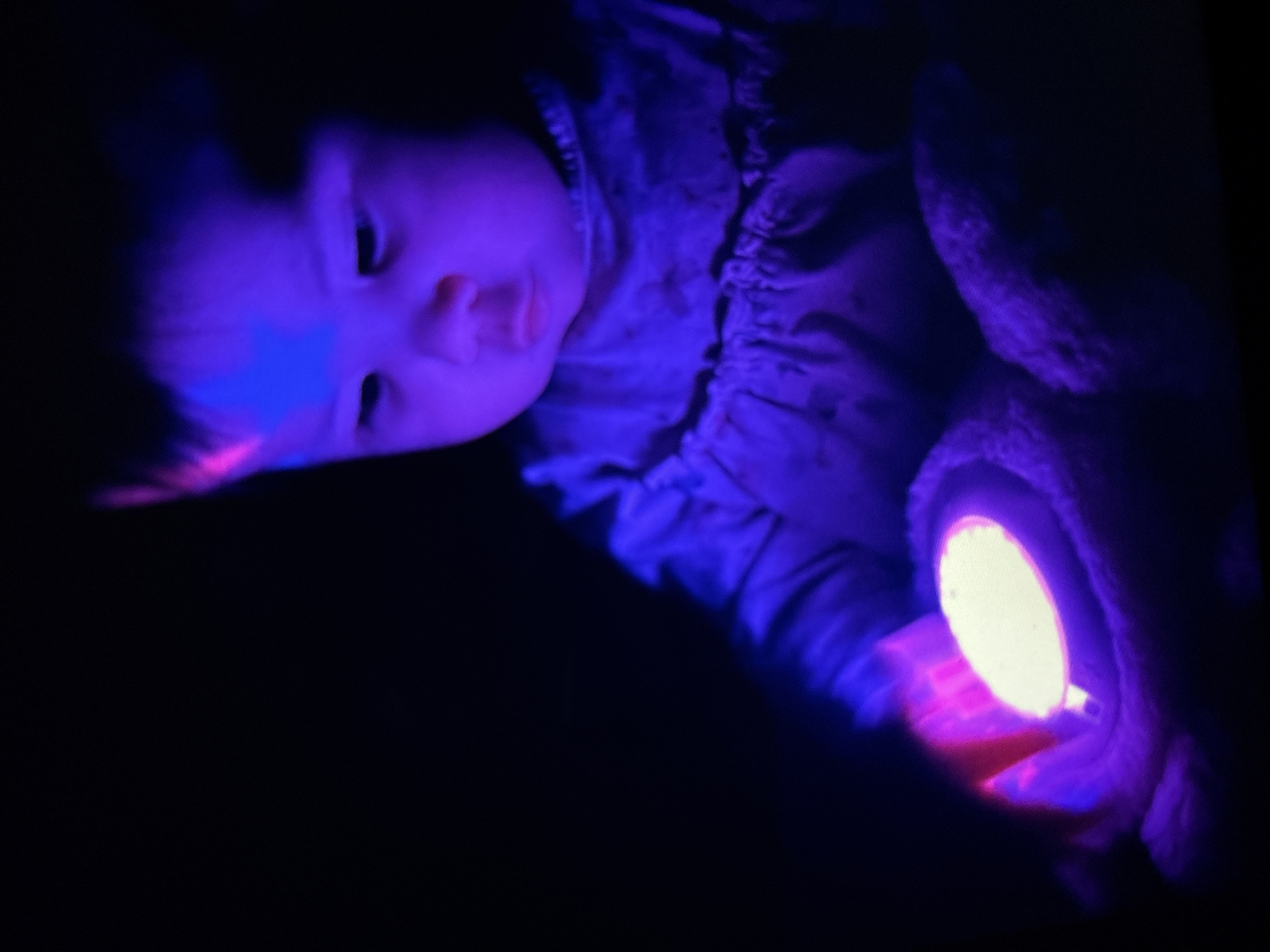 A baby is holding a lit-up object in a darkened environment. The baby is lying down, facing towards the right. The object is emitting a bright light. The location of the photograph is unknown.