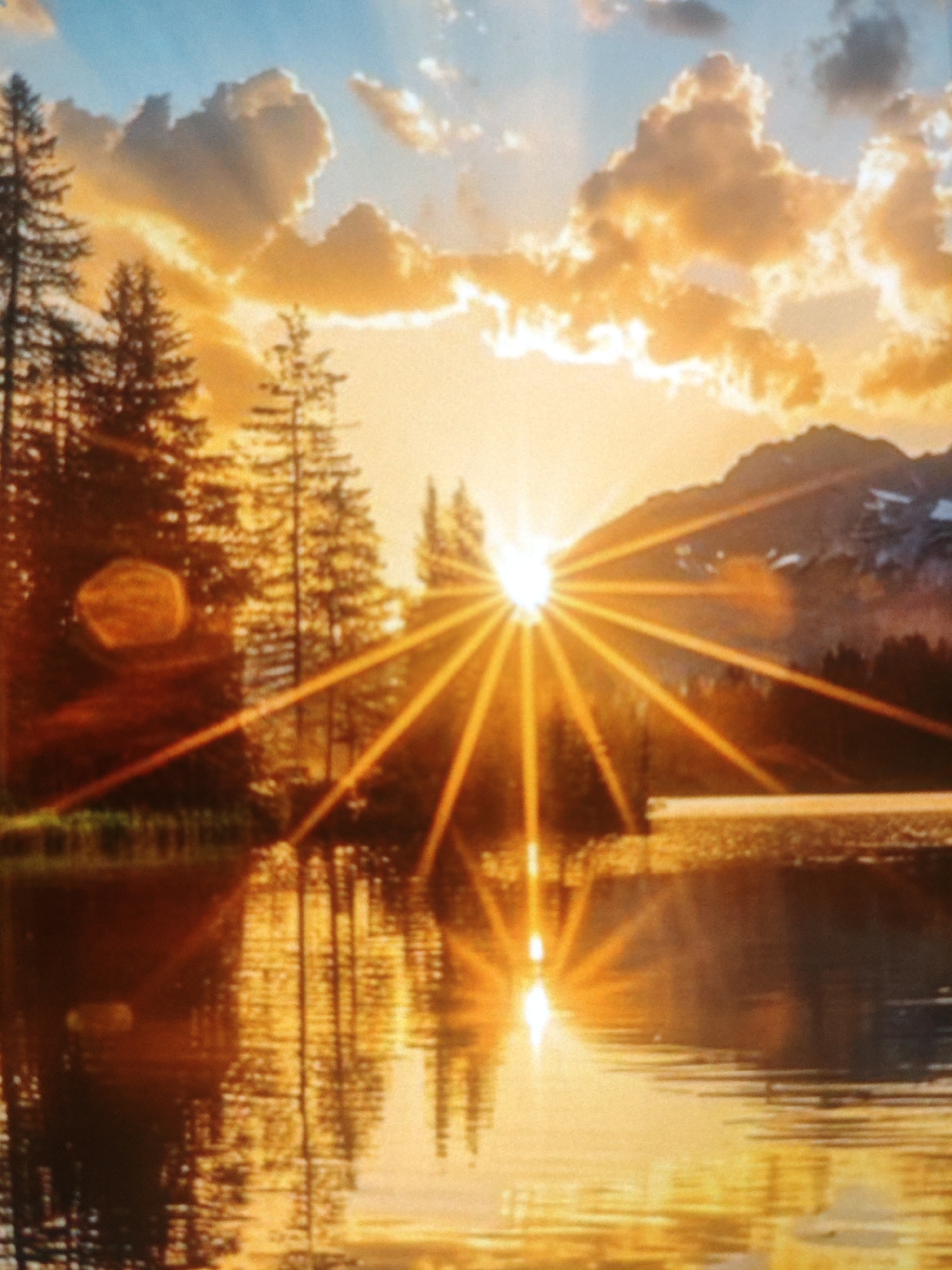 The image depicts a sunset over a body of water, likely a lake or river, with mountains in the background. Tall evergreen trees line the shore on the left side of the frame. The sky is filled with dramatic clouds illuminated by the setting sun, which is casting long rays of light across the scene. The water reflects the vibrant colors of the sky and the surrounding landscape. The overall impression is one of natural beauty and tranquility. There is no information within the image that suggests a specific location in the city of Armavir, Russia.