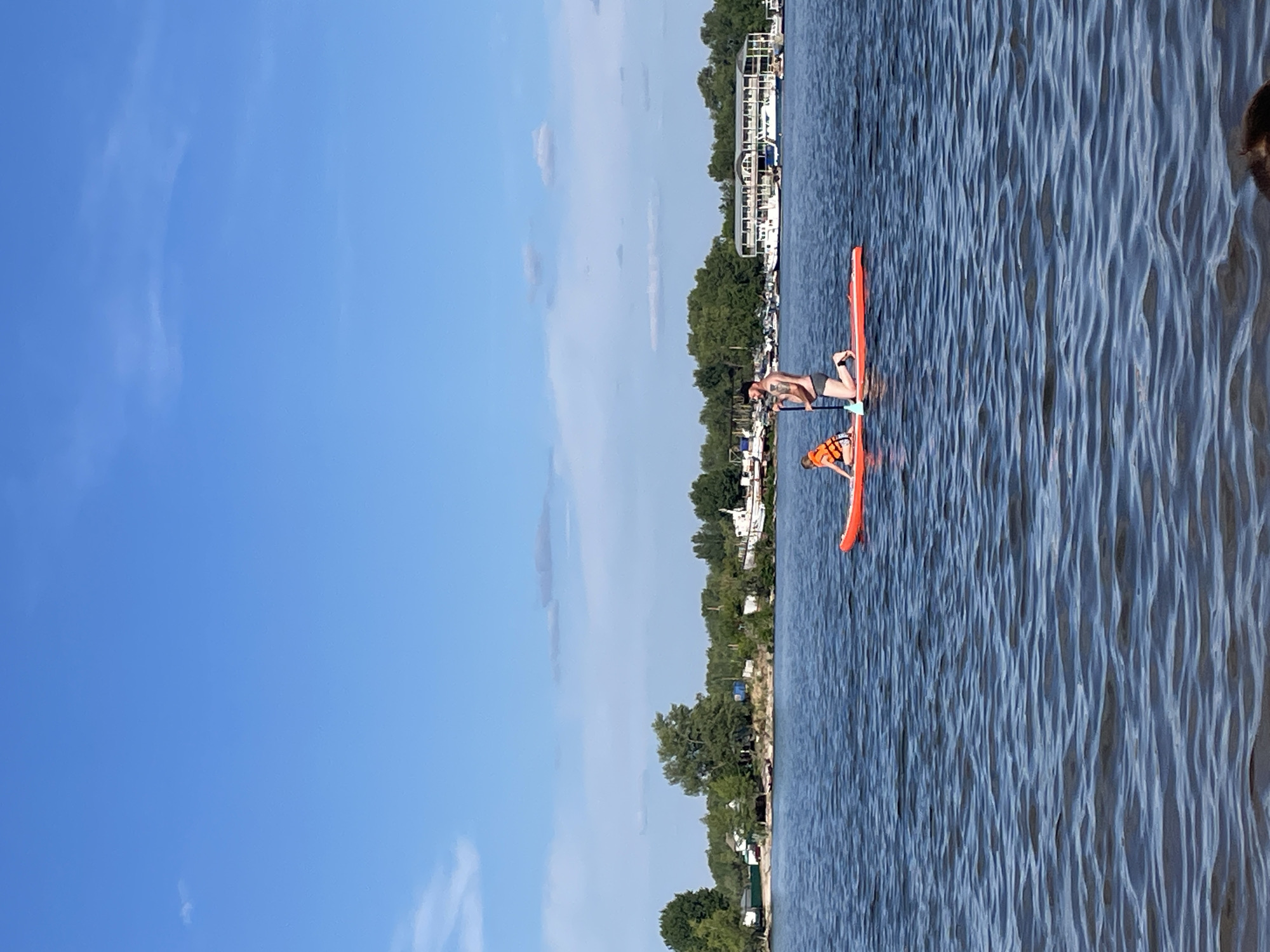 The image shows two people on an orange paddleboard on a body of water. One person, who appears to be an adult, is kneeling on the board and holding a paddle. They are wearing dark shorts and are shirtless. A second person, possibly a child, is sitting on the board in front of the kneeling person and is wearing an orange life jacket. The water is blue and appears to be rippling. In the background, there is a shoreline with trees and some buildings, including a large white boat or structure. The sky is blue with some scattered white clouds. The overall scene suggests a sunny day, likely in the afternoon, given the lighting. The setting appears to be a lake or river with a developed shoreline. The image is oriented vertically, with the sky occupying the left side and the water and shoreline on the right. There is no visible text in the image.
