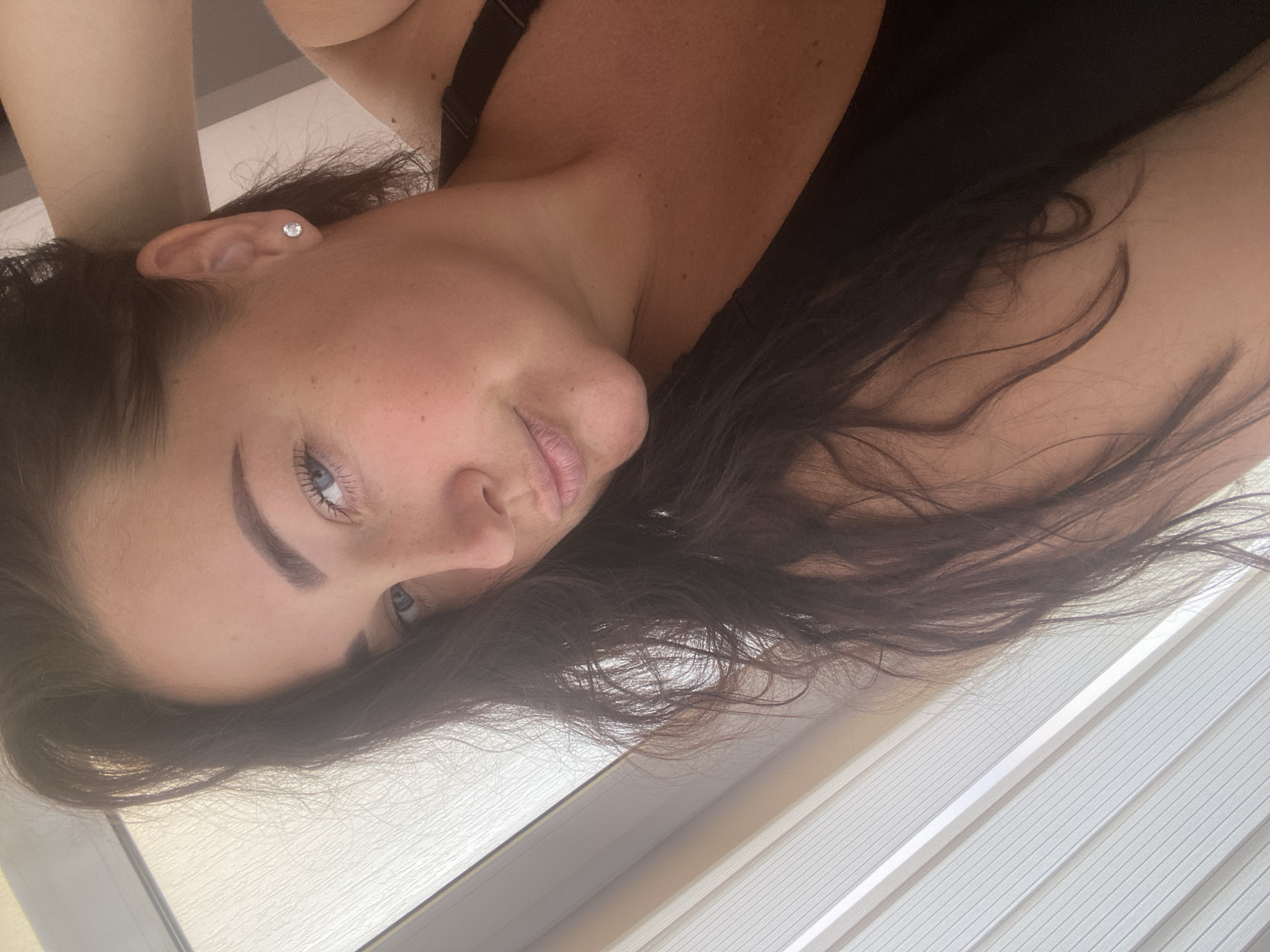 This is a close-up, sideways, selfie-style photograph of a young woman's face. She has dark hair, fair skin with freckles, and blue eyes. She is wearing a dark bra and a diamond stud earring in her left ear. Her head is tilted, and her gaze is directed slightly off-camera. The lighting suggests it could be daytime, with natural light illuminating her face. The background consists of white, slatted blinds. The setting appears to be indoors, possibly in a room with a window. There is no visible text in the image.