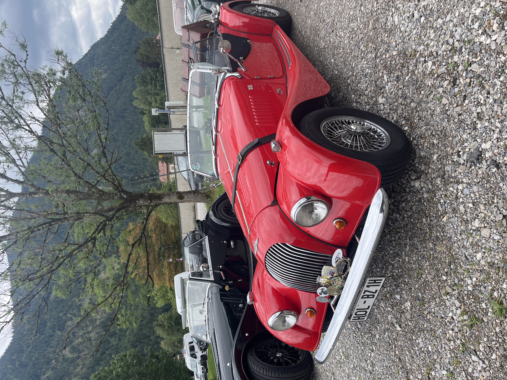 In Ettal, Germany, a red 1960s-era Morgan Plus 4 roadster is parked on a gravel lot in front of a mountain landscape. The car has a chrome grille, wire-spoke wheels, and a luggage rack on the back with a brown leather trunk. A black 1960s-era Morgan Plus 4 roadster is parked to the left of the red car, with its driver's side door open. In the background, a wooded mountain slope rises under a cloudy sky. Several other vehicles, including a white van and a dark-colored sedan, are parked further back in the lot. The scene is likely a parking area for tourists visiting the Ettal Abbey or the surrounding Bavarian Alps.