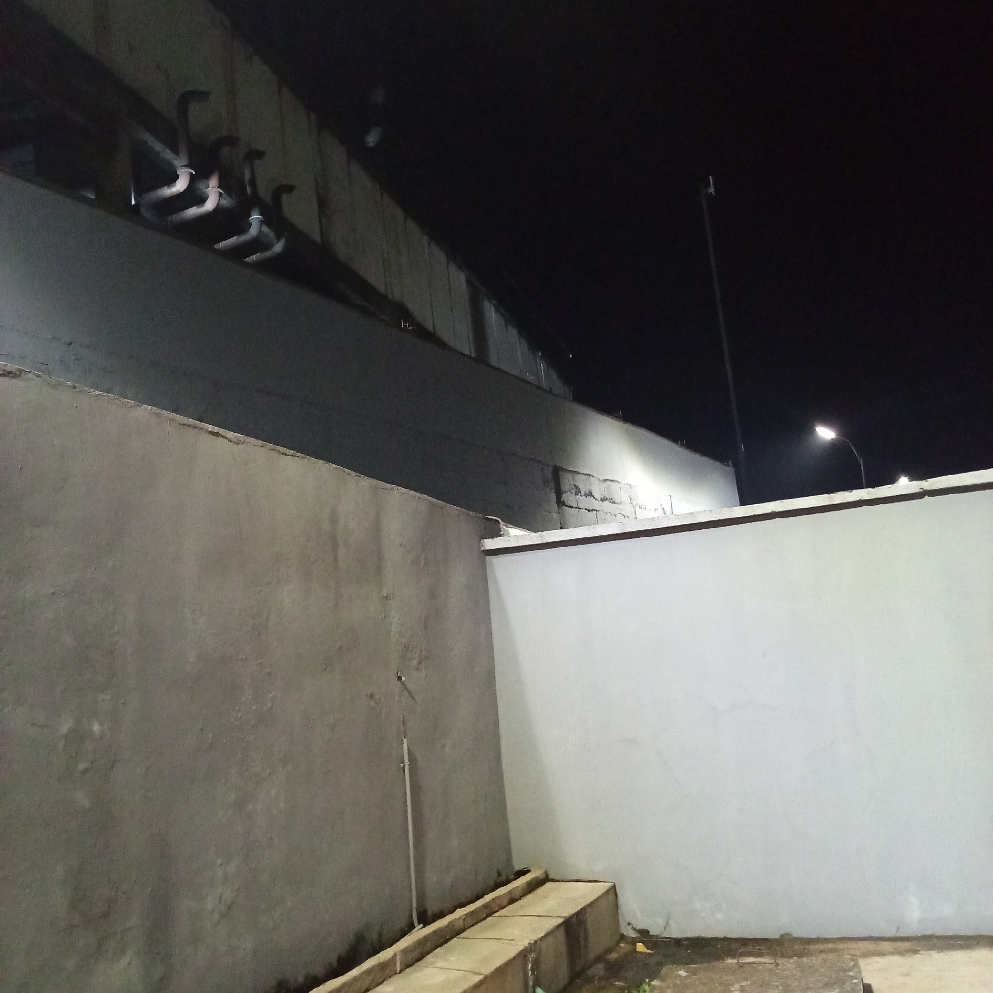 A nighttime exterior shot in Ibadan, Nigeria, depicts a stark, angular environment dominated by concrete walls and a dark, possibly corrugated metal overhang. In the foreground, a rough, grey concrete wall extends across the left of the frame, meeting a lighter, smoother concrete wall that forms the right side. Between these walls, a low concrete structure, possibly a raised platform or step, is visible. Above the grey wall, a section of the overhang juts out, showing several metal pipes or conduits attached to its underside. The background is mostly dark, but a street lamp on a pole is illuminated, casting a bright light on the right-hand wall. The overall impression is one of a functional, perhaps industrial or service area, at night.