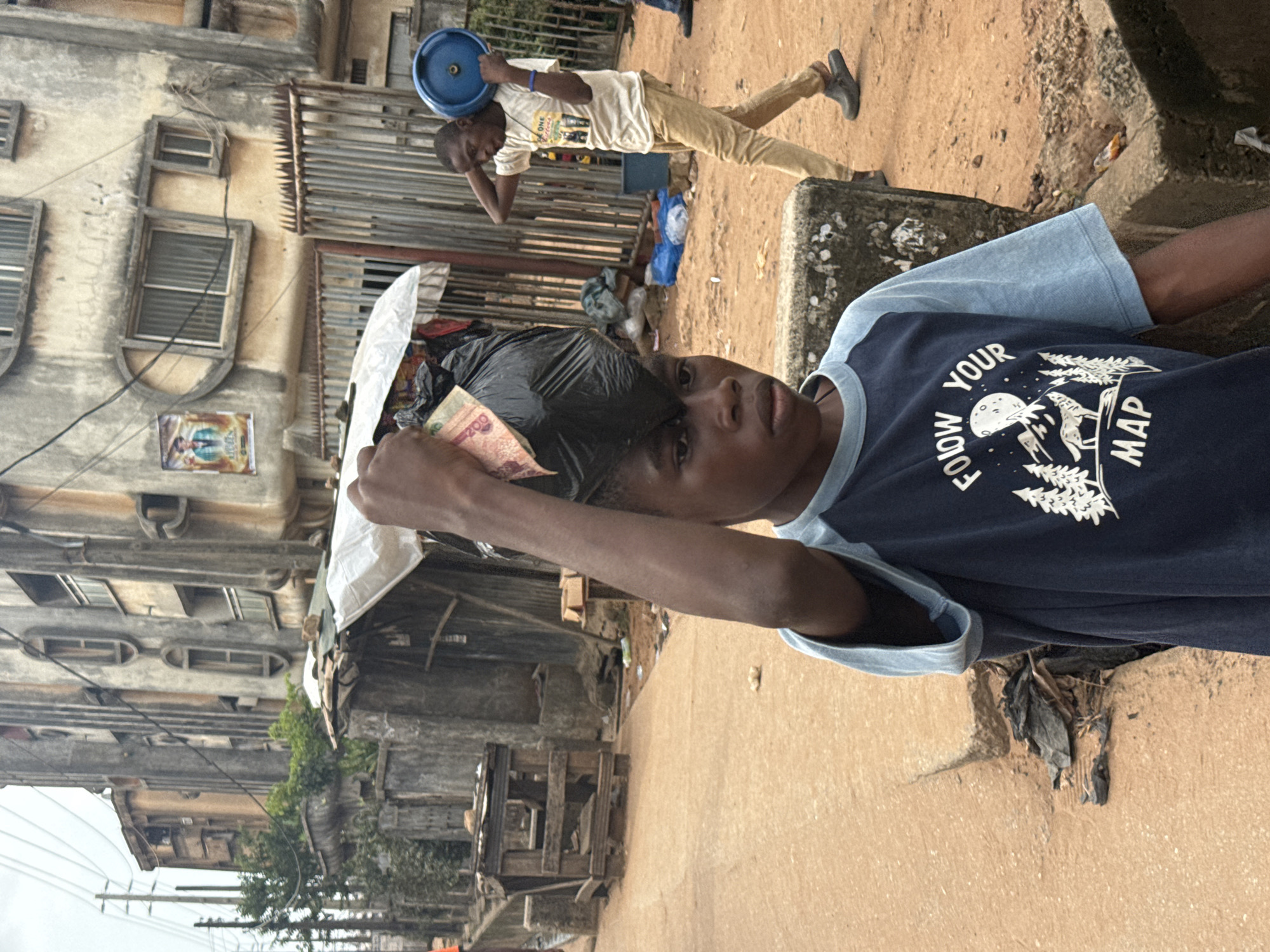 The image shows two young boys in a street scene. In the foreground, a boy wearing a dark blue t-shirt with light blue sleeves and a graphic design that reads "FOLLOW YOUR MAP" is holding a black plastic bag filled with some items, including a banknote that appears to be 200. He is looking towards the camera. In the background, another boy in a light-colored t-shirt and tan pants is walking away from the camera, carrying a blue circular object on his head and holding it with one hand.
The setting appears to be a street in Onitsha, Nigeria, characterized by weathered buildings with multiple windows and balconies, some appearing to be made of concrete. There are also some wooden structures and debris on the ground, suggesting a busy and possibly developing urban environment. The ground is a sandy or dusty color. The time of day seems to be daytime, with overcast or diffused light, possibly indicating a cloudy sky or the early morning/late afternoon. No specific emotions are clearly discernible from the subjects' expressions, but their actions suggest everyday activities in their environment.