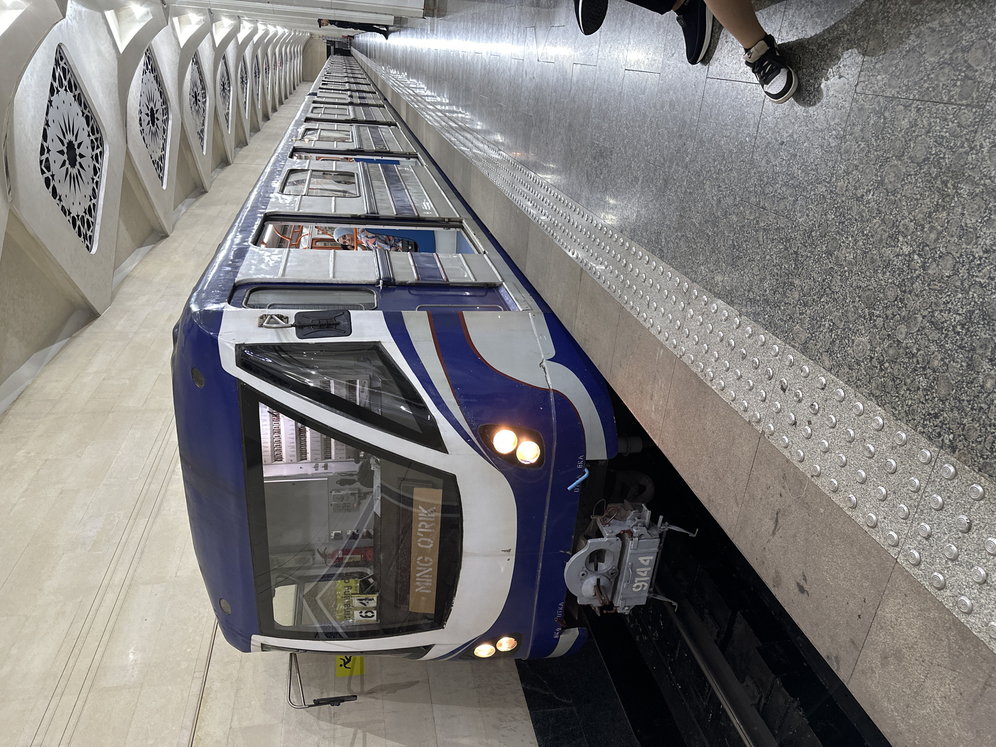 The image shows a blue and white subway train at the "Ming O'rik" station in Tashkent, Uzbekistan. The station features a distinctive arched ceiling with decorative patterns and cream-colored walls. The train's front displays the number "9144" and the destination "MING O'RIK." Several passengers are visible inside the train, including a woman wearing a hijab. The platform is made of granite-like tiles and has a tactile warning strip for visually impaired passengers. Two people are seated on the platform, one adult and one child. The scene is well-lit, suggesting it's an operational subway station.