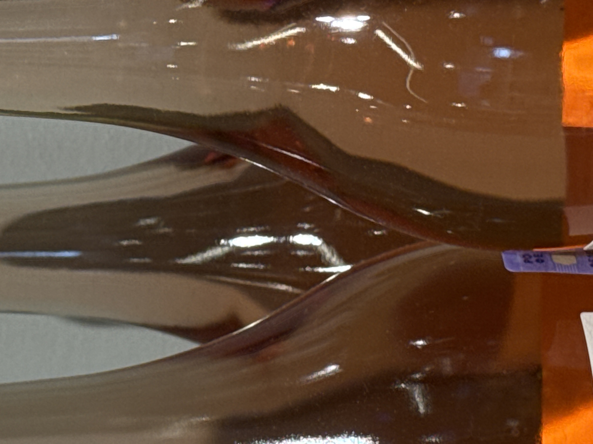 The image shows a close-up, abstract view of amber-colored glass bottles, possibly wine bottles, with a smooth, reflective surface. The light creates highlights and shadows that distort the forms, giving the impression of undulating waves or liquid movement. The bottles appear to be stacked or arranged closely together. In the foreground, a small purple and white label with faint text is visible on one of the bottles. The text on the label appears to be in a sans-serif font and contains characters that might be part of a product code or information. The overall impression is one of a polished, manufactured product. The context of Moscow, Russia, does not provide specific visual cues within this image, as the focus is on the material and form of the bottles. There is no discernible activity or event taking place, and the time of day, weather, or emotions are not conveyed.