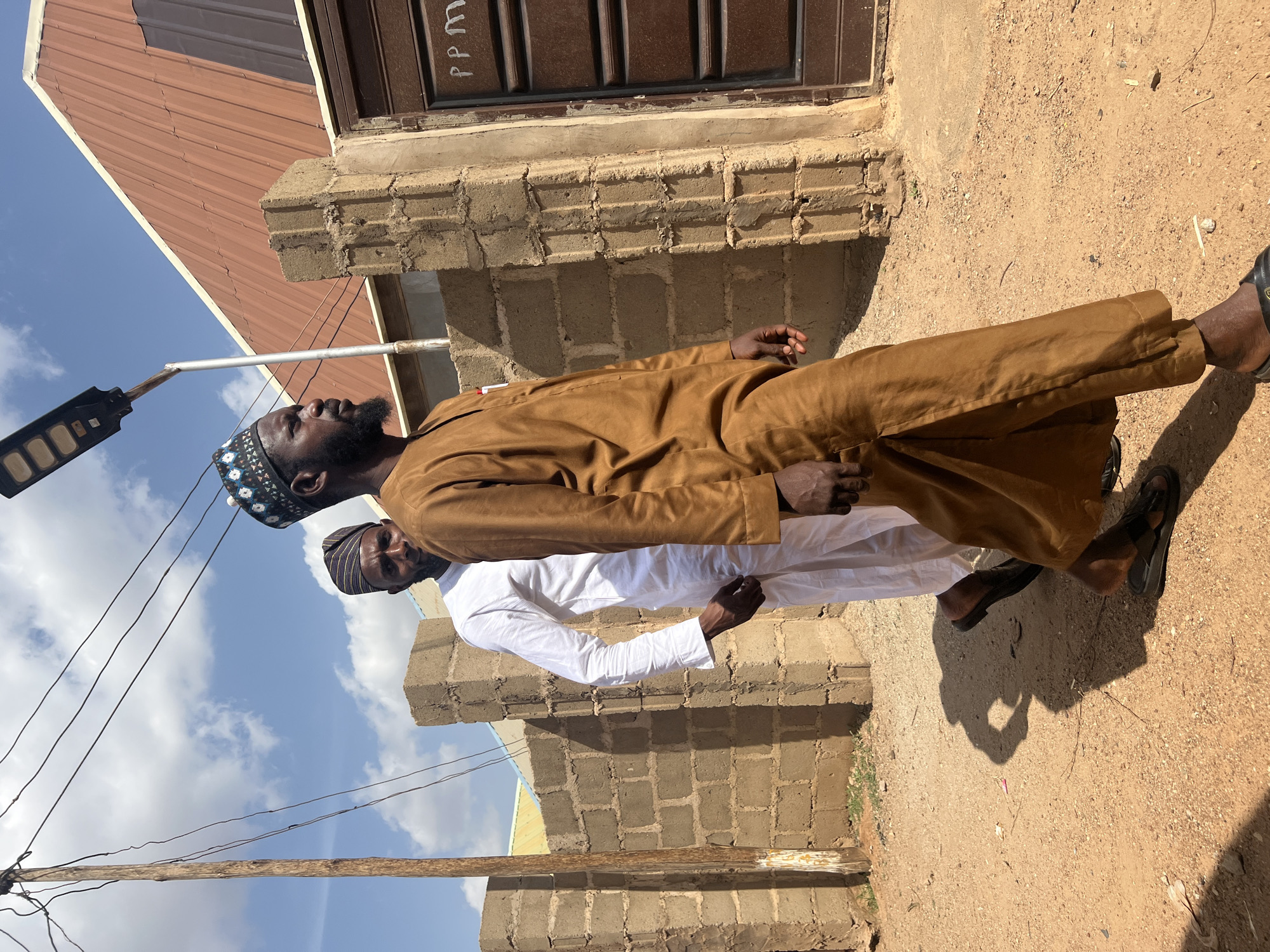 The image shows two men standing outdoors, possibly in the Rigasa area of Nigeria. The man in front wears a brown traditional garment and sandals, with a patterned kufi cap on his head. The second man, partially visible behind him, wears a white garment and a darker kufi cap. A cinder block structure is visible in the background. To the left, there's a building with a reddish-brown roof, and an outdoor light fixture is positioned above the men. The ground appears to be dirt. The sky is partly cloudy.