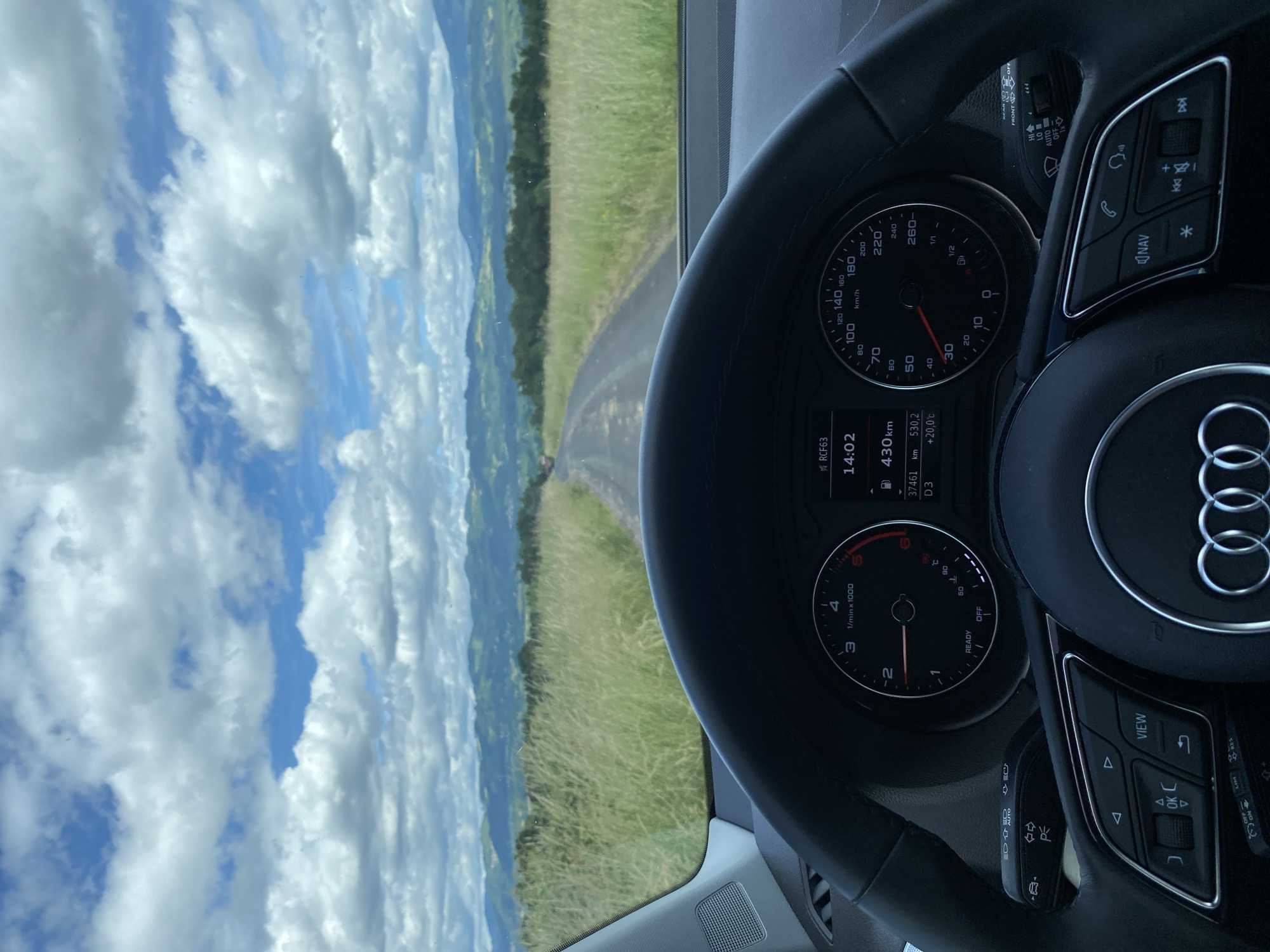 The media file captures a driver's perspective from inside an Audi vehicle, looking out onto a scenic, rural landscape. The image is taken through the windshield, with the car's steering wheel and dashboard occupying the foreground.
In the interior, a black leather steering wheel with the Audi logo is prominent on the right. Various buttons are visible on the steering wheel, including controls for navigation ("NAV"), phone, volume, track skipping, and menu options ("VIEW", "OK", "RES", "LIM"). The dashboard features two main dials: a speedometer showing values up to 260 km/h, and a tachometer up to 6 x 1000 rpm. Both needles are at zero, indicating the car is stationary. A digital display between the dials shows several pieces of information: "RCF63", the time "14:02", remaining range as "430km", a trip meter reading "530.2km", and an outside temperature of "+20.0°C". Other readings include "37461" (likely total odometer) and "D3" (drive mode). Stalks behind the steering wheel show controls for wipers (HI, LO, AUTO, OFF) and lights (AUTO, OFF, ON, with symbols for headlights and fog lights).
Beyond the car, a narrow, winding asphalt road curves through lush green grassy fields. In the distance, rolling hills or mountains are visible under a partially cloudy sky, with patches of clear blue. The overall setting suggests a pleasant journey through the countryside, possibly in the Courpière region of France, given the contextual information. The time of day is early afternoon, and the weather is mild and bright.