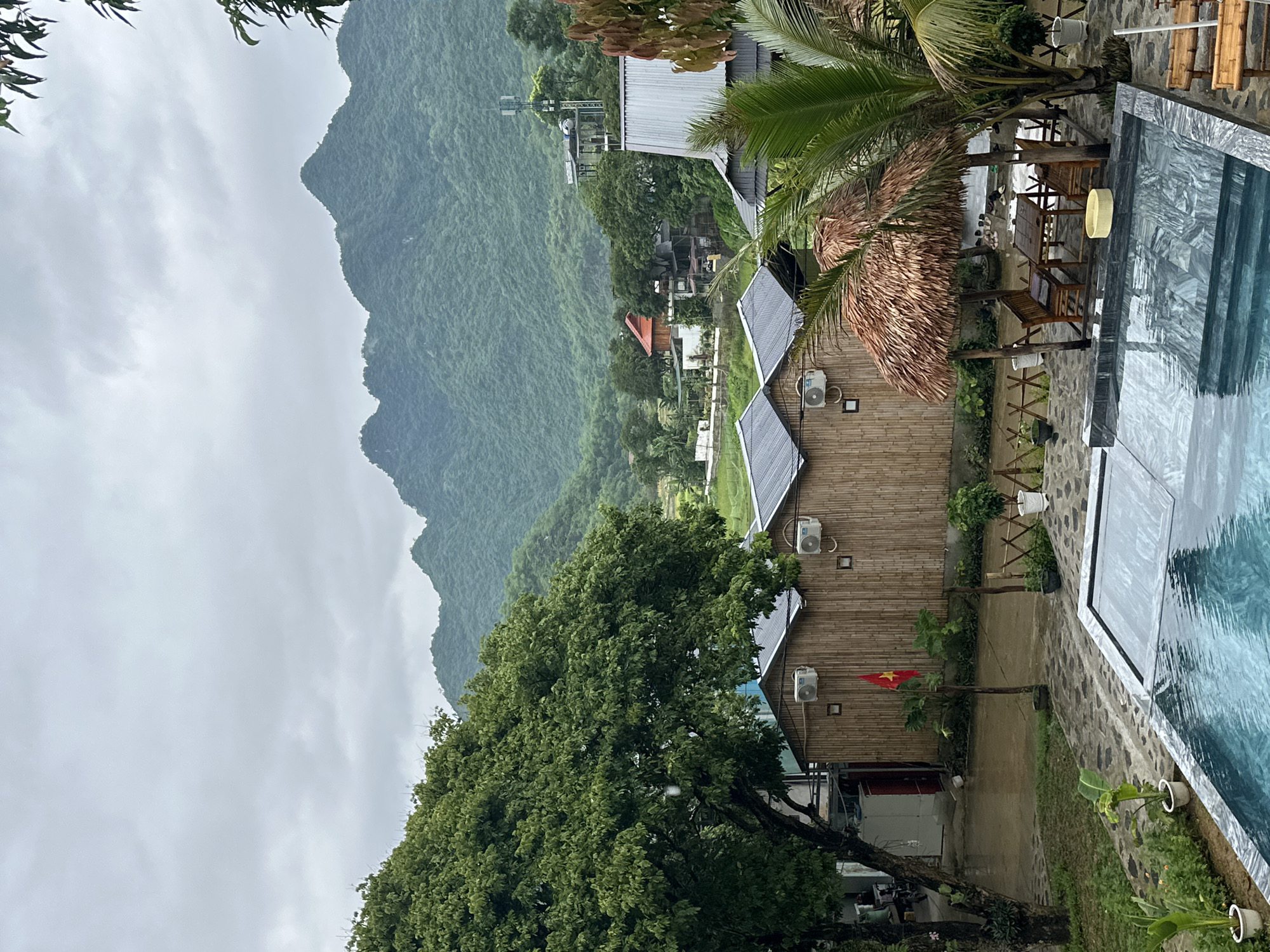 The image depicts a serene, cloudy day at a resort in Xóm Khán, Vietnam. In the foreground, a rectangular swimming pool with a reflective, dark-tiled edge is partially covered by a transparent tarp. Adjacent to the pool is an outdoor seating area with a thatched-roof gazebo, furnished with a wooden table and four chairs. A palm tree stands to the right of the gazebo.
Behind the seating area, a row of three identical bamboo-clad bungalows are visible, each equipped with an air conditioning unit. A Vietnamese flag on a pole stands in front of the central bungalow. Further in the background, lush green mountains dominate the landscape under a heavily overcast sky. Scattered buildings and trees are nestled between the bungalows and the mountains. The ground surrounding the pool and bungalows is paved with grey stone tiles and patches of wet earth, suggesting recent rain. The overall scene is tranquil and natural, indicative of a tropical retreat.