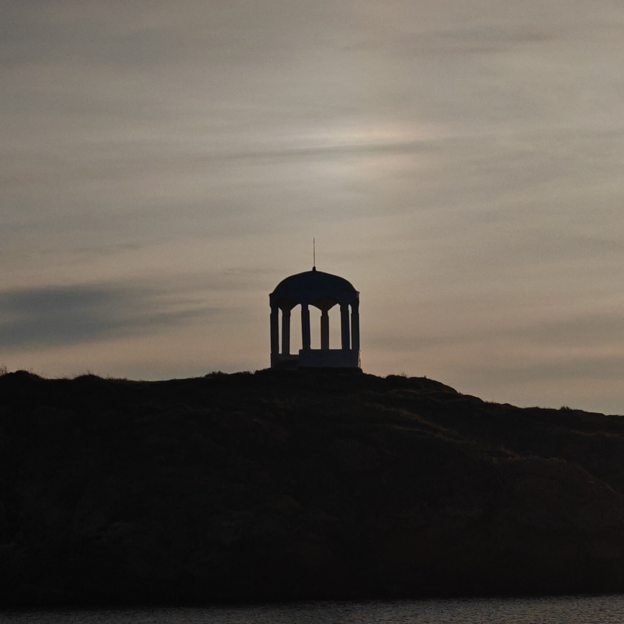 A silhouette of a structure resembling a gazebo sits atop a dark hill or mound against a pale, overcast sky. The structure is the Temple-Rotunda located in Мысовское сельское поселение, Ukraine. The hill descends to a body of water in the foreground. The scene is characterized by low light, with the subjects appearing in shadow. The sky is light gray with subtle variations suggesting cloud cover.