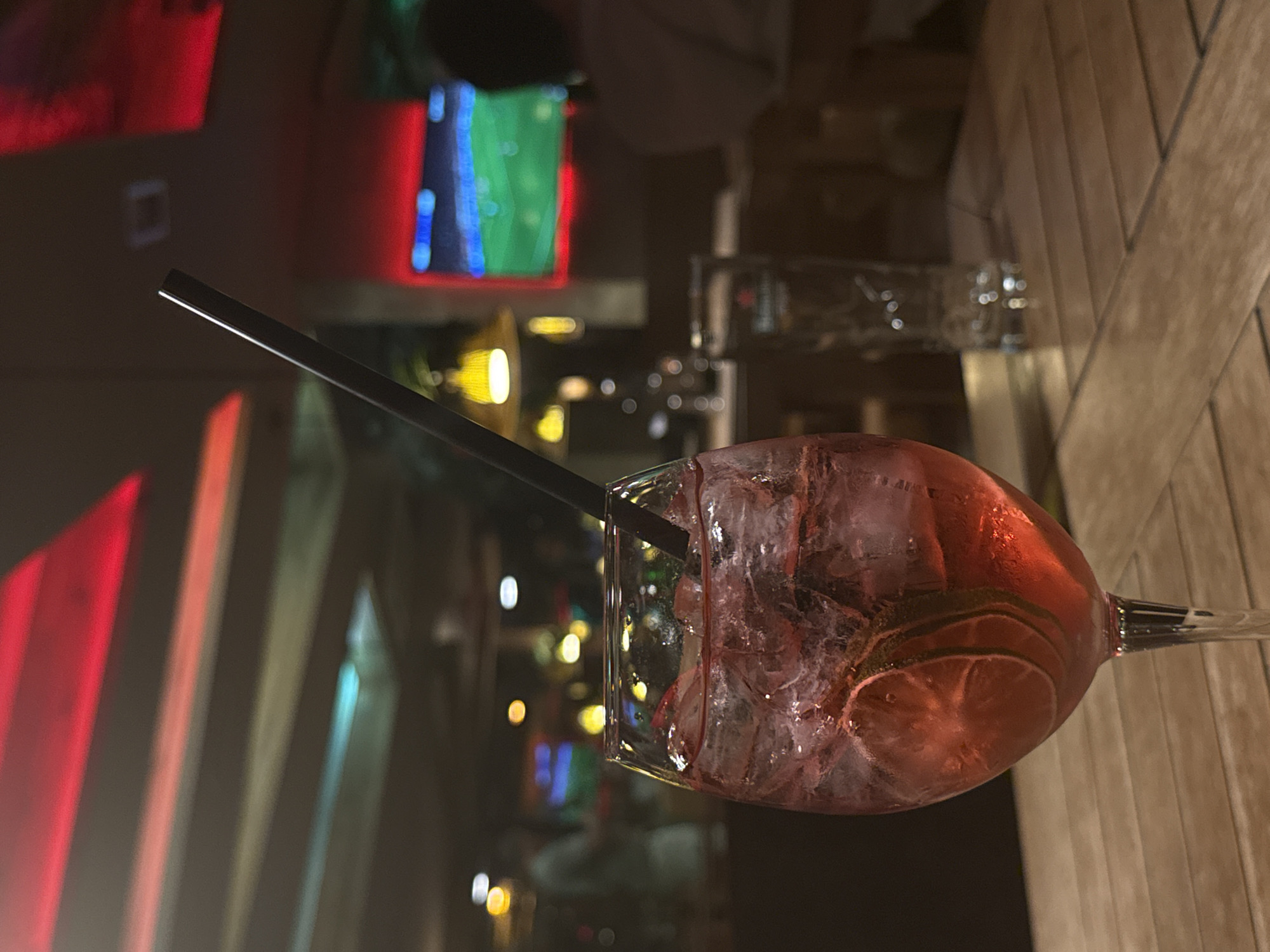 The image shows a cocktail with ice and lime slices in a wine glass, with a black straw. Another glass containing a clear liquid is visible in the background. The cocktail is placed on a wooden table with a vertical grain pattern. The background shows a blurred cityscape with colorful lights, suggesting an urban environment, possibly at night. Based on the available information, determining the specific location in Sofia, Bulgaria is not possible.