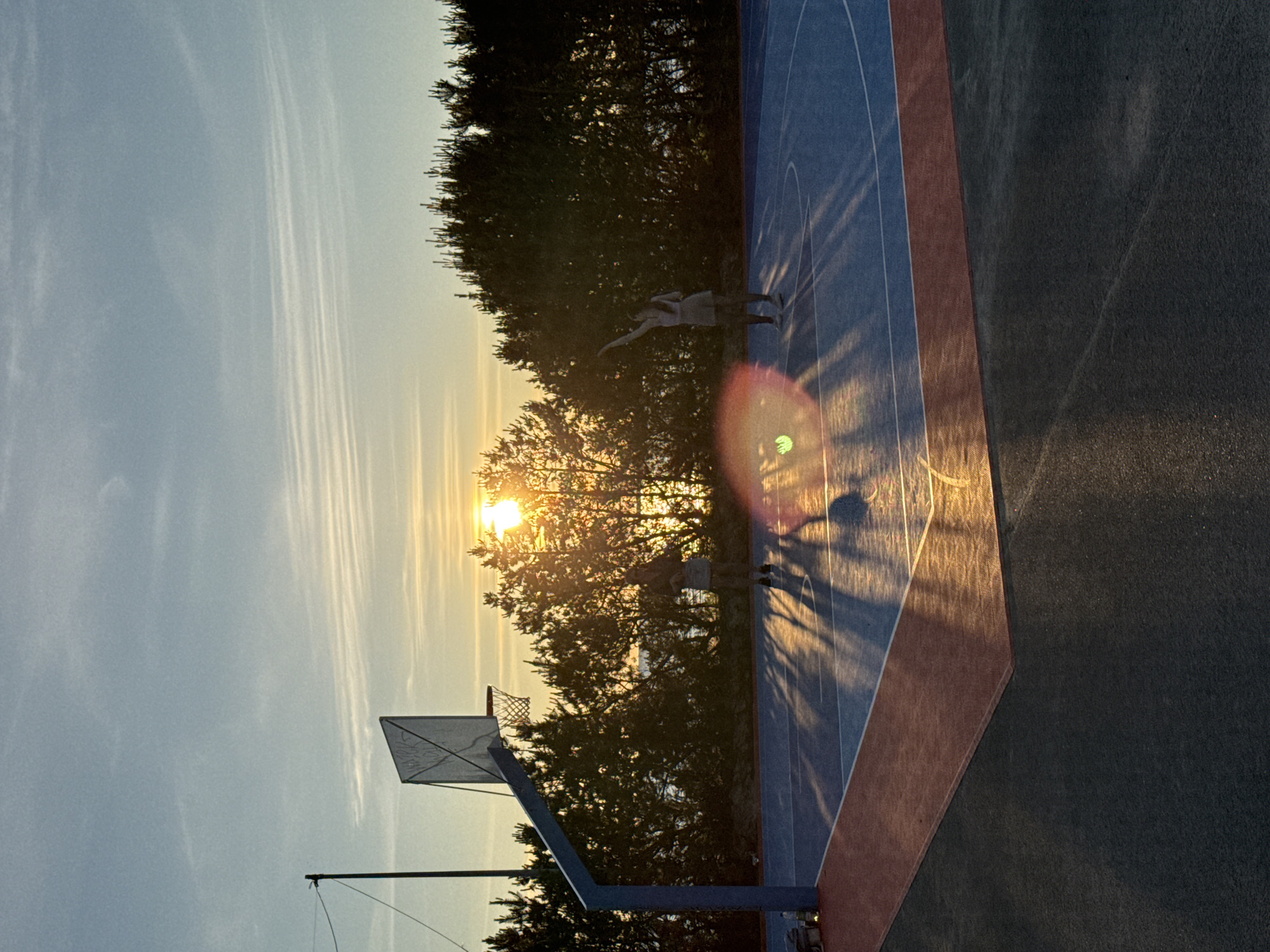 This is a vertical, medium shot photograph of a basketball court at sunset. The court itself is painted blue with white lines and a red border. A basketball hoop with a white backboard is visible on the left side of the frame. Two silhouetted figures are playing basketball on the court. One figure is on the left side, near the hoop, while the other is on the right, in the process of shooting. The setting sun is low on the horizon, behind a line of dark green trees, casting long shadows across the court. There is a lens flare in the lower right portion of the image, with a circular green glow and rays emanating from it. The sky is a pale blue with wispy white clouds. The scene appears to be outdoors, possibly in a park or recreational area. No specific landmarks or identifying features of Tallinn, Estonia are visible in the image.