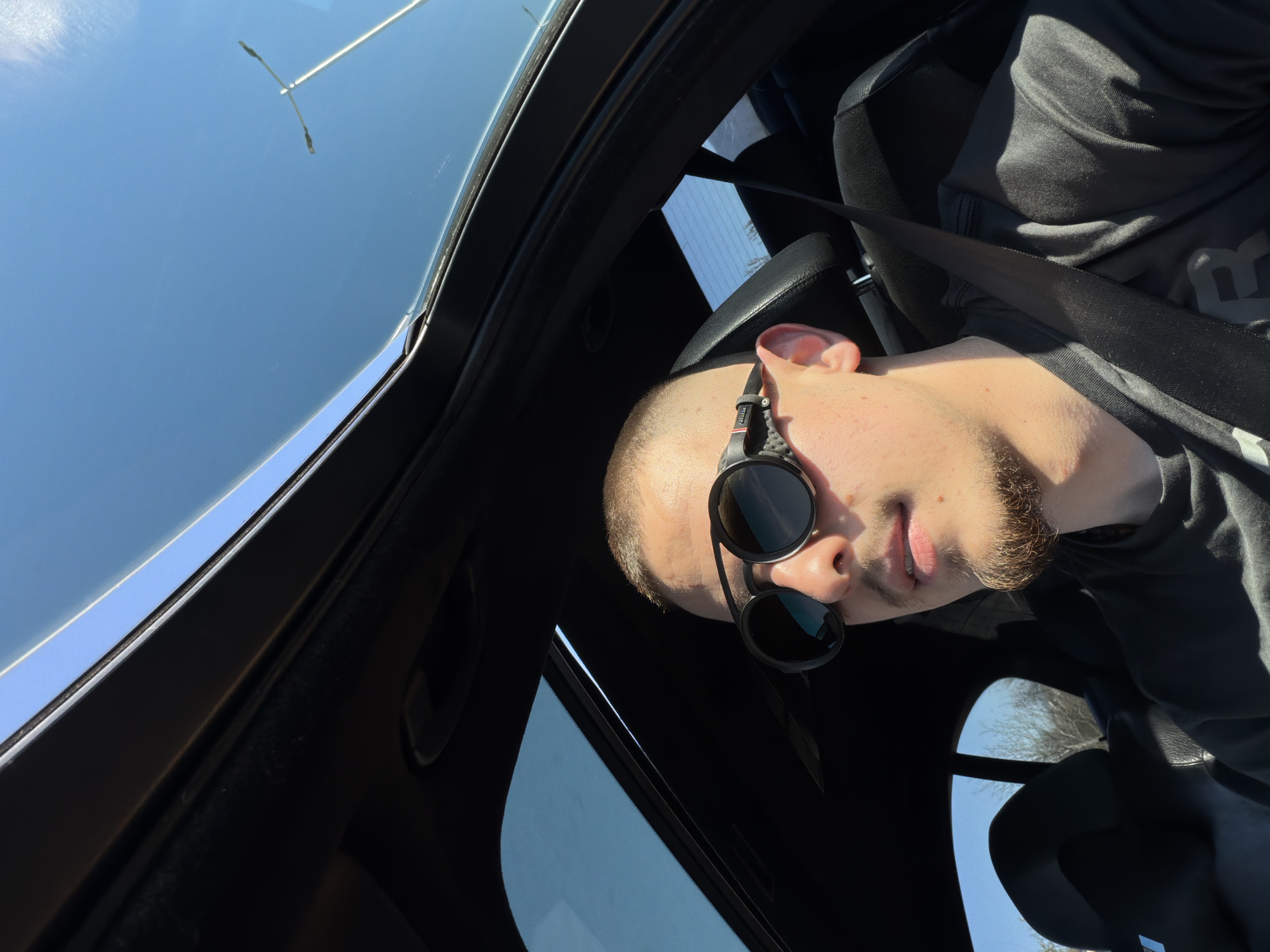 A man wearing sunglasses and a black shirt is seated in a black car, secured with a seatbelt. The car appears to have a sunroof, and the bright sky is visible through it, along with a tall street lamp. Based on the architectural style of the car's interior, it is not possible to determine the precise location in Plovdiv, Bulgaria without further context or identifying landmarks. The angle of the photograph suggests it was taken from inside the vehicle looking upwards.