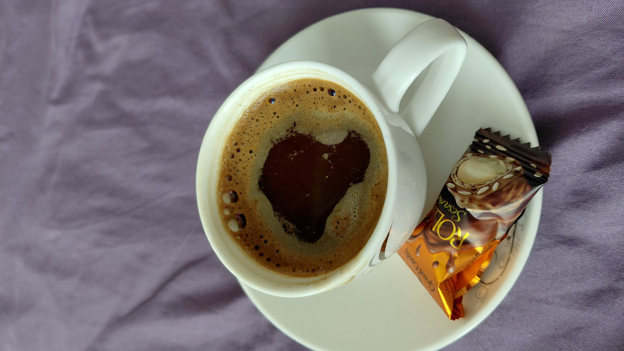 A close-up, top-down shot captures a white ceramic cup filled with dark coffee, complete with a foamy crema, resting on a white saucer. The saucer is adorned with a subtle, swirling pattern around the rim. Positioned in front of the cup on the saucer is a single, individually wrapped chocolate candy with a gold and brown wrapper. The wrapper features text in a stylized font, including "Grand Candy" and "POLO Sesami," and displays an image of the candy inside, showing nuts and what appears to be sesame seeds. The entire arrangement is set against a backdrop of a textured, lavender-colored fabric, likely a tablecloth or bedspread. There are no people visible in the image. The scene appears to be indoors, possibly in a domestic setting. No specific location in the city of Abovyan, Armenia can be determined from the visual information provided.