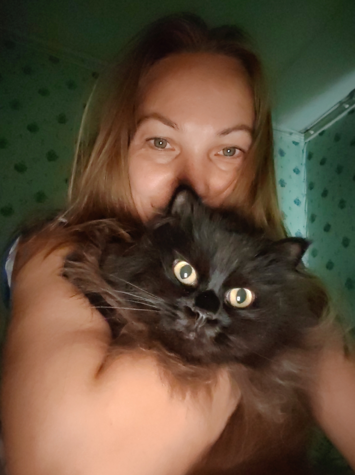 A person is holding a black cat in their arms. The person's face is partially visible, with their eyes looking at the camera and a smile on their face. The cat's face is also visible, with its eyes wide open and looking at the camera. The background appears to be a room with green wallpaper. The image is a selfie taken by the person. There is no information in the image to suggest it is located in Чухлэм, Russia.