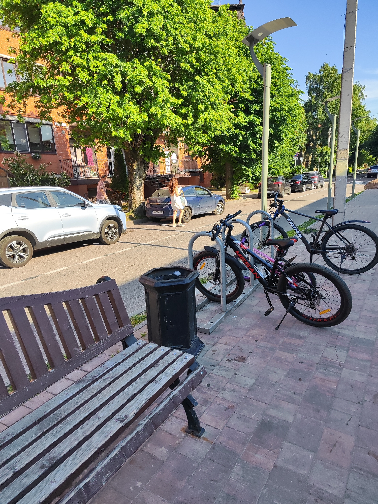 The image depicts a street scene in Pionersky, Russia. In the foreground, a weathered wooden park bench faces a sidewalk paved with bricks. Next to the bench is a dark-colored trash receptacle. Behind the bench, a line of bicycles is parked at a bike rack. The bicycles are of various colors, including black, pink, and purple.
The background reveals a street with several cars parked along the side and driving on the road. A light-colored SUV is parked in the lane, while other dark-colored cars are visible further down. On the left side of the street, a brick building with balconies and large windows is situated behind a large, leafy green tree. On the right side of the street, modern lampposts are visible, and more trees line the sidewalk.
Two people are visible in the middle ground. One person, wearing a white dress, stands near the parked SUV. Another person, wearing a striped shirt and shorts, is walking towards the building. The overall atmosphere suggests a sunny day, likely in the late afternoon or early evening given the quality of the light. The scene appears to be in an urban or suburban residential area. No visible text is legible in the image.