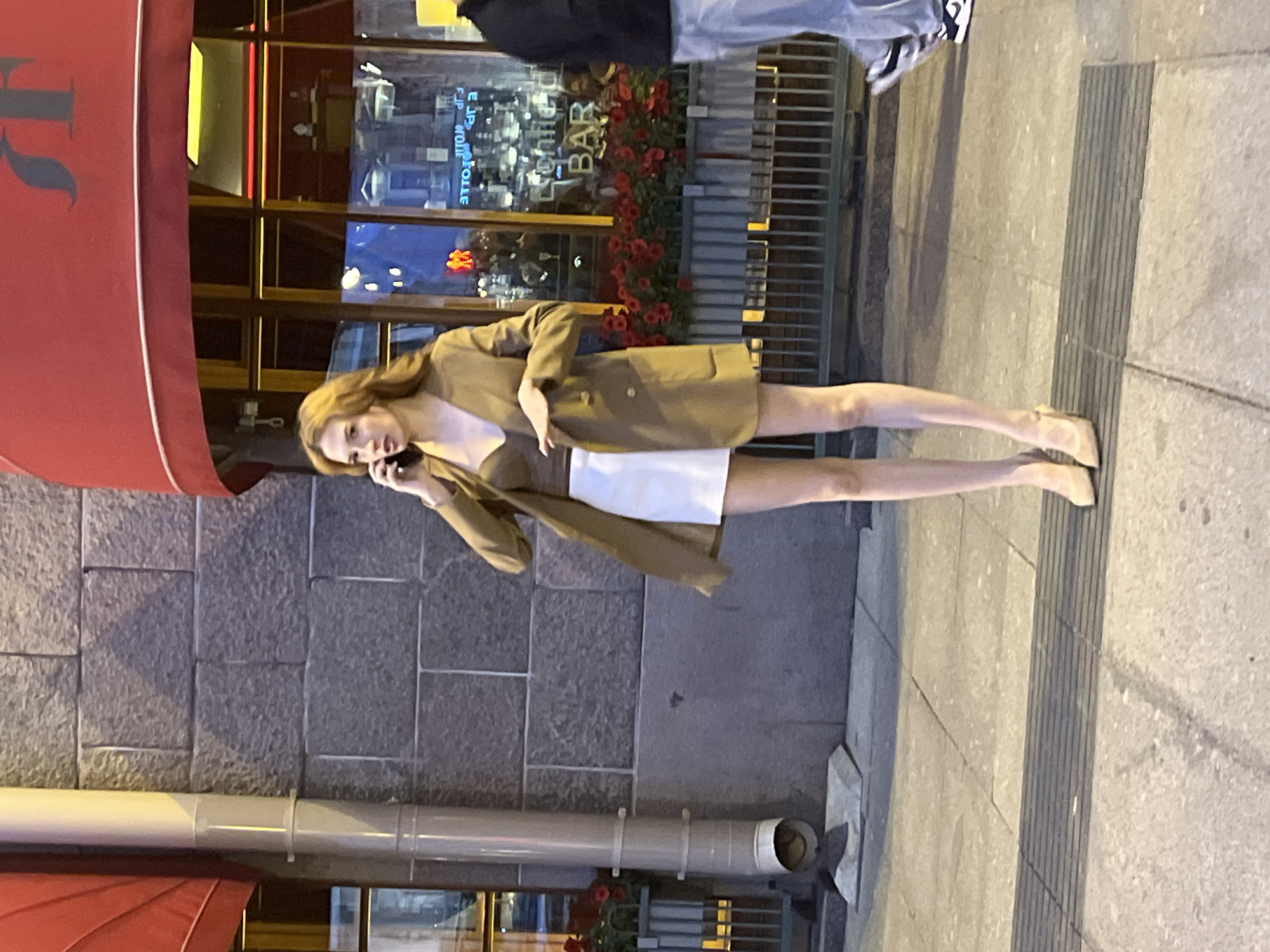 A young woman with auburn hair is standing on a sidewalk in front of a building with a red awning and large windows. She is wearing a brown blazer over a brown top and a white mini-skirt, along with beige high-heeled shoes. She is holding a mobile phone to her ear and appears to be engaged in a conversation. The building behind her has a stone facade and planters with red flowers. The street level of the building is a bar with illuminated signs. The scene appears to be a street in Saint Petersburg, Russia, possibly Nevsky Prospekt, given the architecture and atmosphere. A portion of another person's legs, wearing blue jeans and sneakers, is visible on the right side of the frame, walking away from the camera. The lighting suggests it is evening or dusk.
