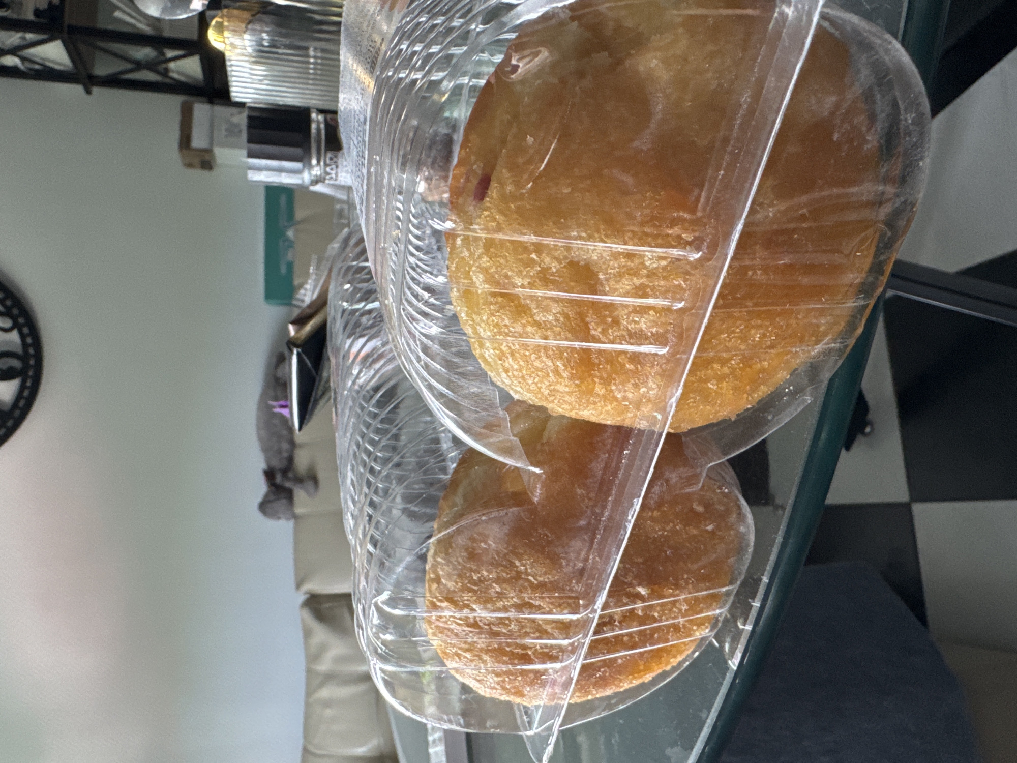 The image shows two round, golden-brown pastries, possibly doughnuts or fritters, encased in clear plastic packaging. The pastries have a slightly textured surface. The packaging itself appears to be a transparent plastic container with horizontal ridging. They are placed on a glass surface, which reflects the checkered pattern of a floor or tabletop in the background. To the left, part of a black and white circular object, possibly a clock face, is visible. In the background, there are hints of household items, including what appears to be a shelf unit with some boxes and a metallic object with a light source. A blurred figure or object, perhaps a stuffed animal, is partially visible behind the pastries on the left. The overall setting seems to be an indoor environment, possibly a kitchen or dining area. There is no visible text in the image. The lighting suggests it might be daytime, but there are no strong indicators of specific time or weather.