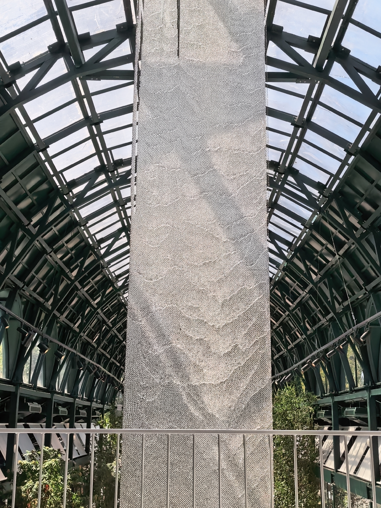 The image shows the interior of the "Gulliver" shopping mall in Moscow, Russia, specifically focusing on the atrium. The viewpoint is from a balcony or upper level, looking towards a large, textured column that dominates the center of the frame. The column is covered in a light-colored, mesh-like material.
Above and to the sides of the column, the architecture of the mall's roof is visible. It features a complex structure of dark green metal beams and supports, with translucent panels that allow natural light to filter through. This creates a geometric, almost skeletal pattern against the sky.
In the foreground, a silver metal railing runs horizontally across the bottom of the image, separating the viewer from the space below. Beyond the railing, lush green trees and foliage can be seen, indicating the presence of indoor landscaping or a botanical garden within the mall. Spotlights are also visible mounted on a dark structure in the background, suggesting the area is illuminated for retail or events.
The overall impression is one of a modern, well-lit, and architecturally interesting interior space, designed to provide a pleasant shopping and recreational environment.