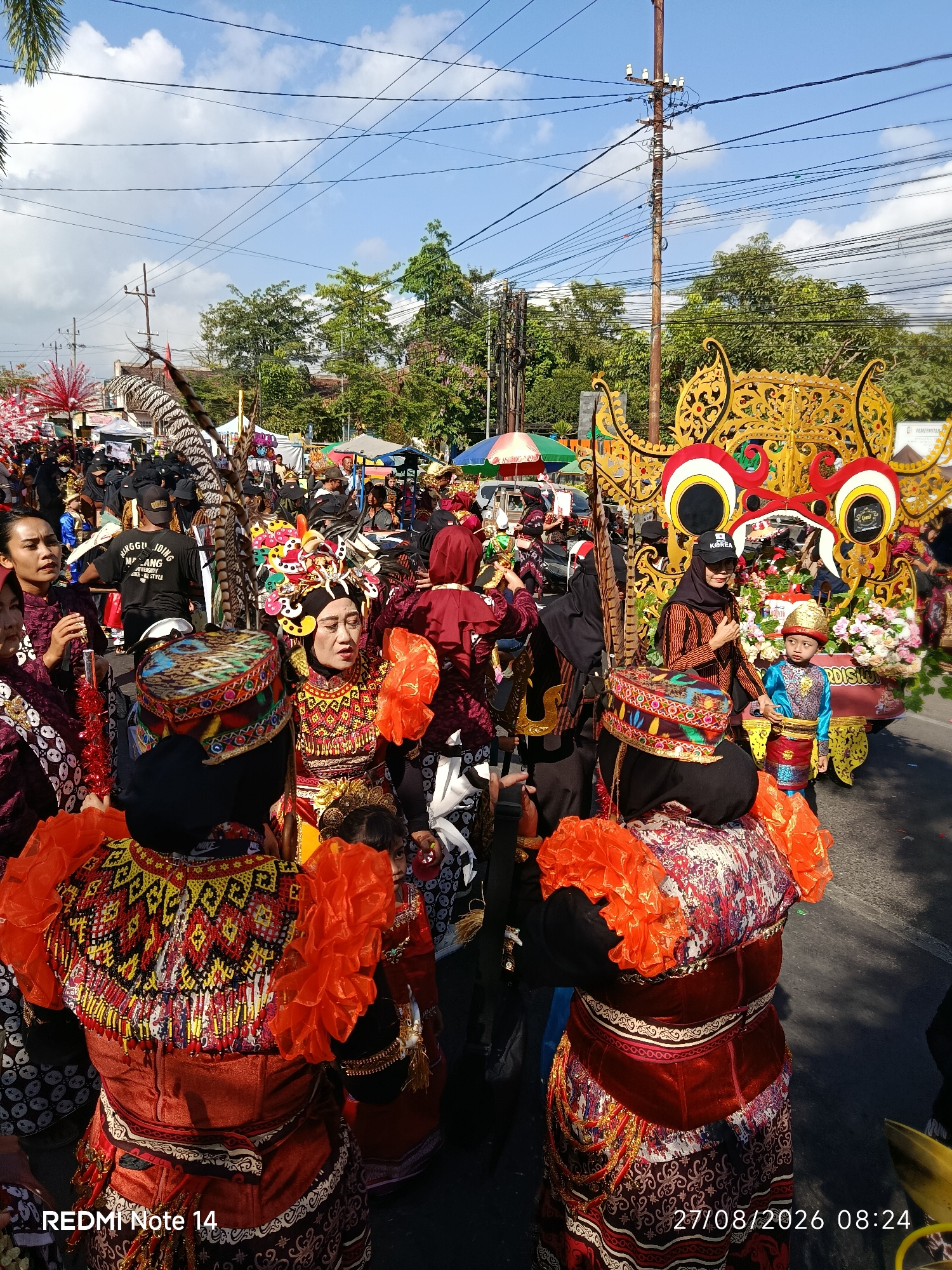 This image captures a vibrant cultural procession or parade in the city of Kalangbret, Indonesia. The scene is bustling with people, many adorned in traditional, colorful attire. In the foreground, several individuals are dressed in ornate, embroidered outfits featuring prominent orange ruffled sleeves and intricately patterned headwear. One woman in the center has elaborate gold and red head ornamentation. Behind them, a large, golden, dragon-like float or structure, decorated with red and white accents, is visible, suggesting a significant celebratory event. A young boy in a blue and gold traditional outfit stands near the float. The background reveals a street lined with trees, tents or stalls, and power lines, indicating an urban or semi-urban setting. The sky is blue with scattered clouds, and the lighting suggests it is daytime, likely late morning or early afternoon. The overall atmosphere appears festive and communal.