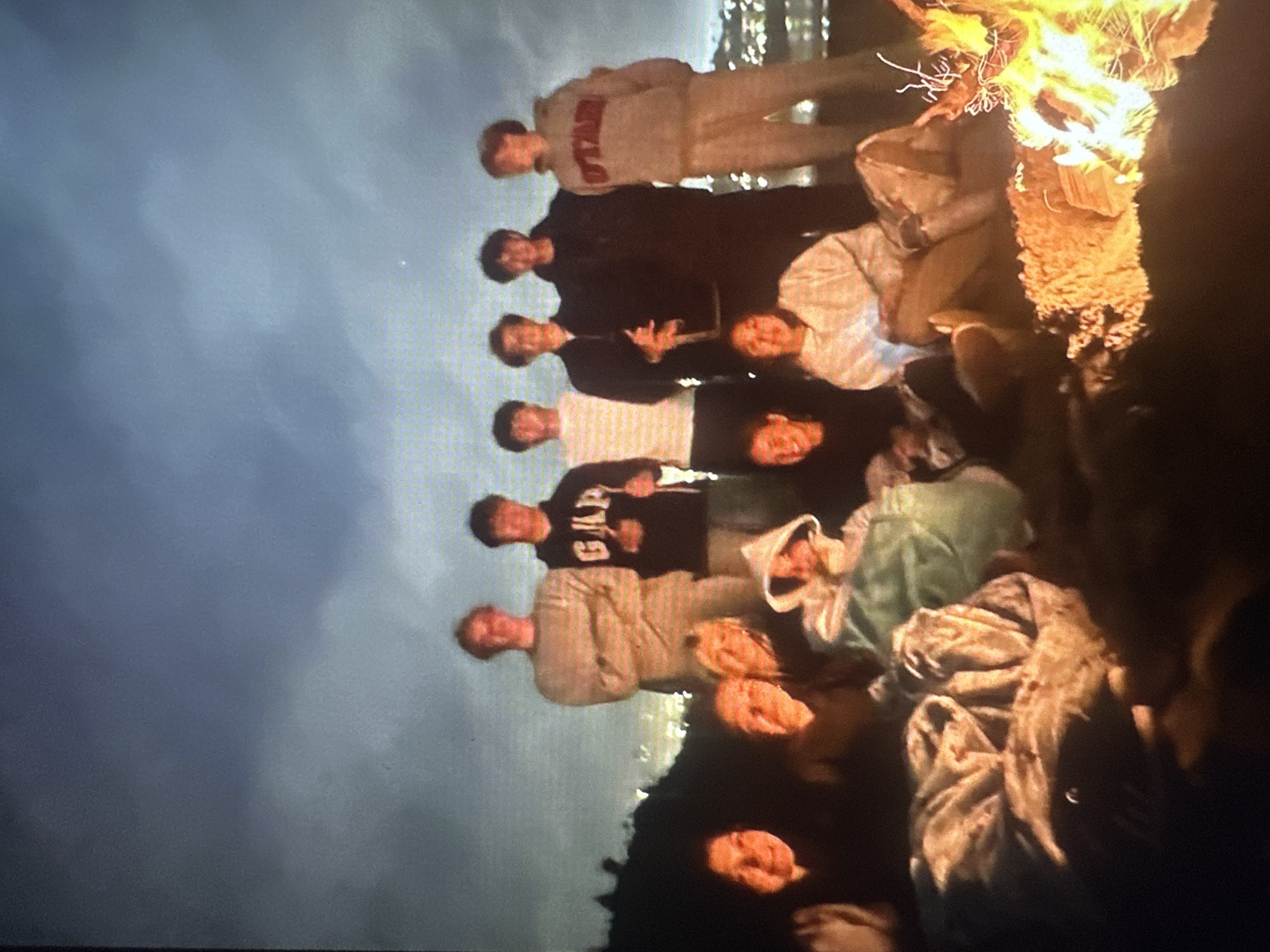 The image shows a group of ten young people gathered around a bonfire at night, with a city skyline visible in the background. They are seated and standing, some are looking at the camera, others are engaged in conversation. The fire is burning brightly, casting a warm glow on their faces. The sky is dark and cloudy. The city lights suggest an urban environment, potentially Tallinn, Estonia, though this cannot be definitively confirmed from the image alone.