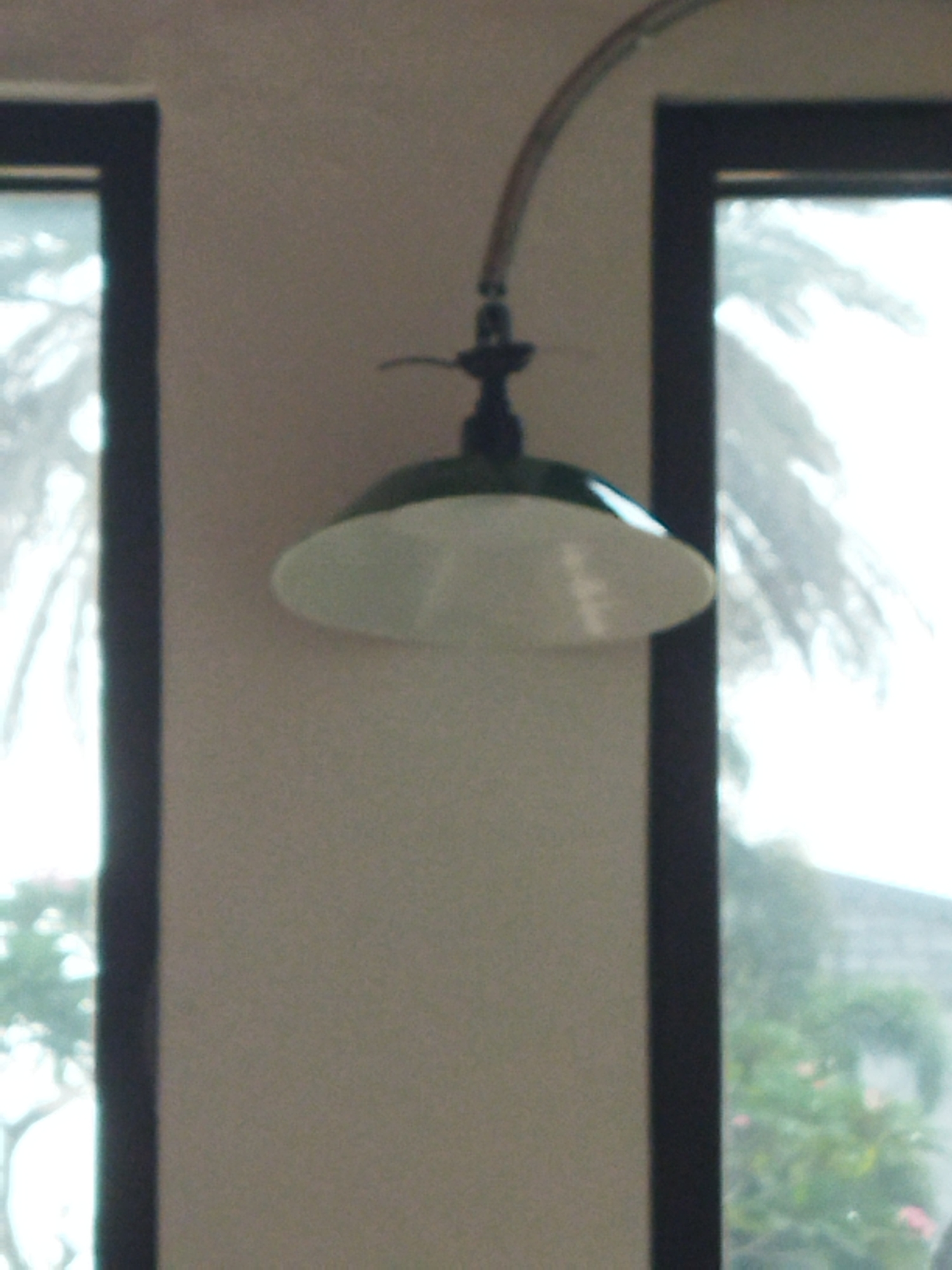 The image shows a vintage-style pendant light fixture hanging from a curved metal arm. The light fixture has a dark green top and a white, saucer-shaped shade. It is positioned in front of a wall with two dark-framed windows. Through the windows, palm trees and foliage are visible, suggesting an outdoor setting with lush vegetation. The overall scene appears to be indoors looking out at a tropical or semi-tropical environment. The image is slightly grainy and out of focus, contributing to a nostalgic or hazy aesthetic. The location is stated to be in Suka Sari, Indonesia.
