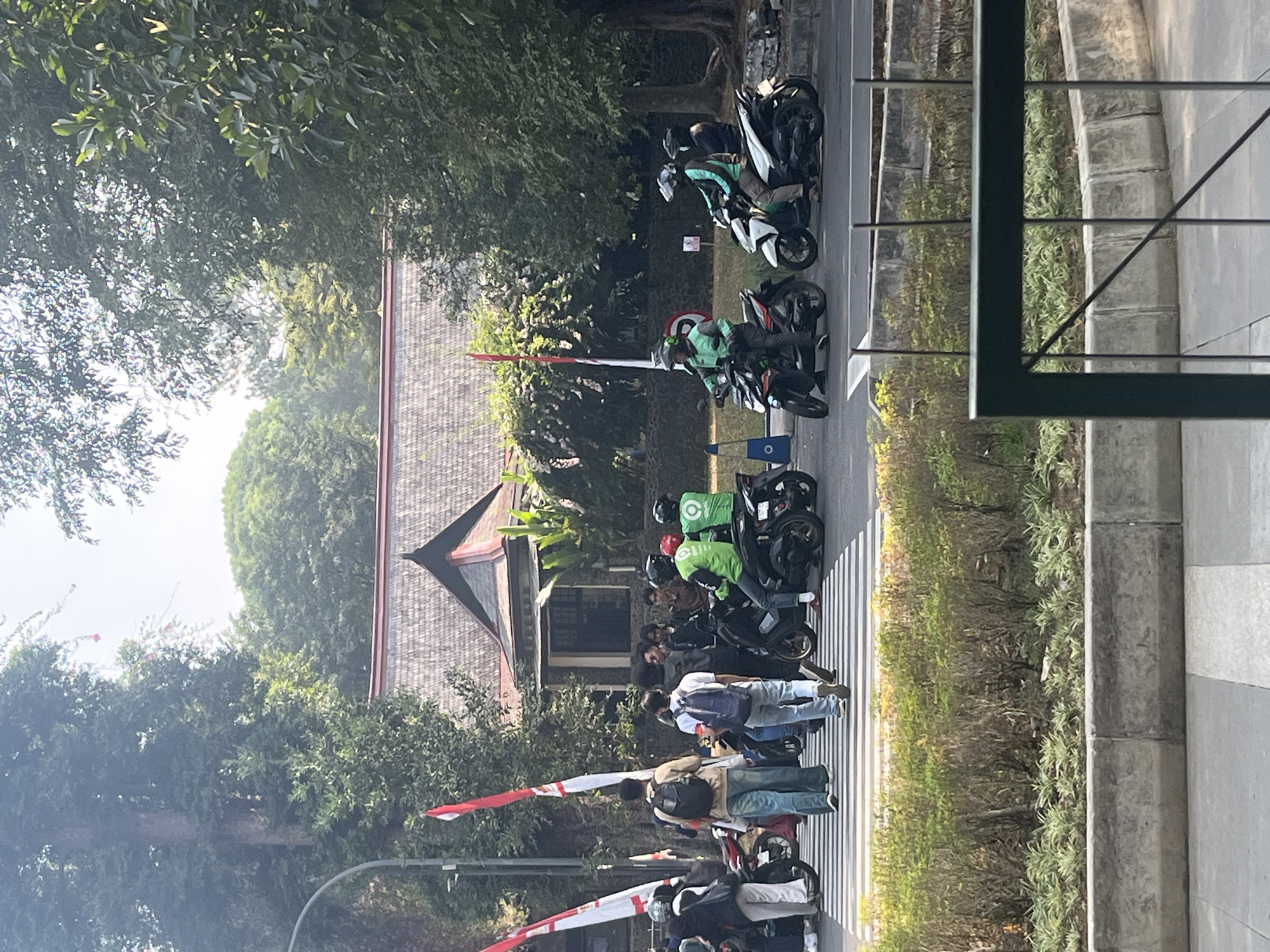 A street scene unfolds in Bandung City, Indonesia, on a bright day. A pedestrian crossing is occupied by several people and motorcycles. Two individuals, one wearing a backpack, walk across the zebra stripes. To their right, three men on motorcycles are stopped. Two of them wear green jackets with the "Gojek" logo, indicating they are likely ride-sharing drivers. A third man behind them wears a black jacket. Further to the right, two more individuals on motorcycles are also stopped, their faces obscured by helmets.
In the background, a building with a distinctive tiled roof and stone accents stands amidst lush greenery. Tall trees line the street, their branches casting dappled shadows. Two red and white Indonesian flags on poles are visible on the left side of the image, suggesting a national celebration or event. The overall impression is one of urban activity, with transportation, pedestrians, and buildings coexisting under a clear sky.