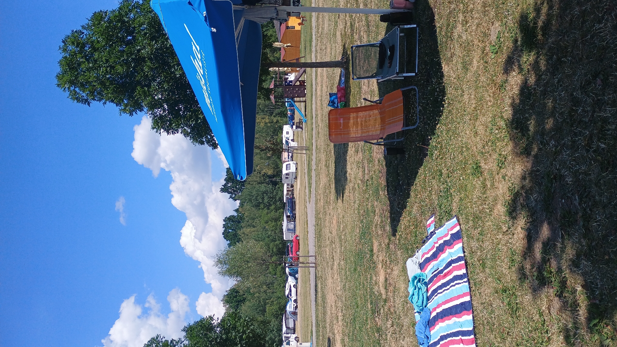 The image depicts a campground scene on a sunny day, likely in Sokolov, Czechia, given the provided context. In the foreground, a striped beach blanket with towels is laid out on dry grass. Two lounge chairs, one orange and one dark grey, are positioned under a large blue umbrella. The umbrella has some text, partially visible. In the mid-ground, a playground with a slide and climbing structure is visible. Several tents, caravans, and vehicles, including a red car, are parked in the background. A line of trees forms a backdrop behind the campground. The sky is blue with scattered white clouds.