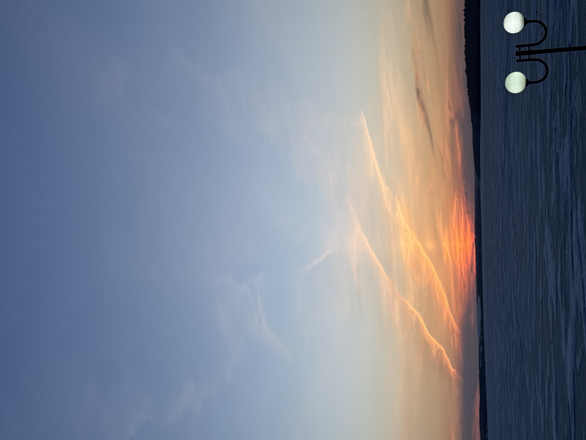 The image shows a sunset over a body of water, likely a lake or river, in Votkinsk, Russia. The sky transitions from blue to orange and red near the horizon, with some wispy clouds visible. The shoreline is dark, with trees silhouetted against the sky. A street lamp with two spherical lights is visible on the right side of the image. The water appears calm with some textures, potentially indicating ice formation.
