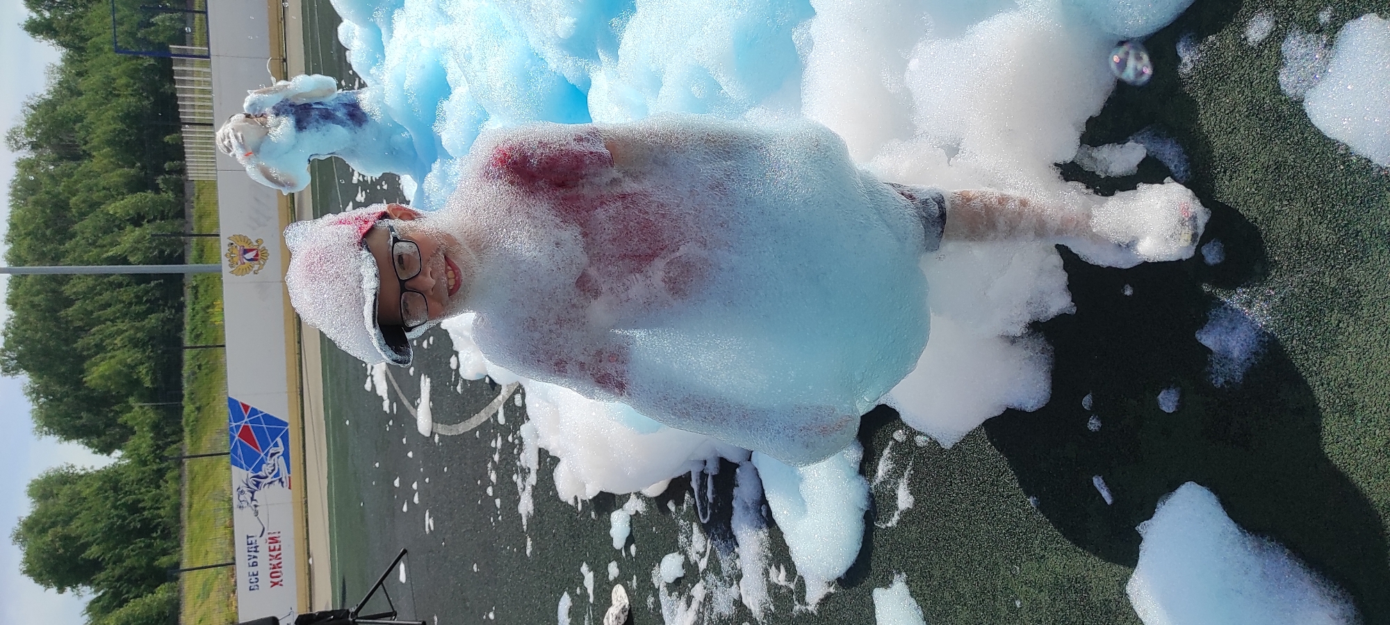 This media file shows a young person partially submerged in a large amount of blue and white foam. The person is wearing glasses, a red shirt, and a dark cap. They are smiling and appear to be enjoying themselves. Their body is mostly covered by the foam, with only their face, arms, and legs visible in parts. The foam appears to be the result of a foam party or event.
The scene is outdoors, with a grassy field and a fence visible in the background. There is a white wall with some logos and text on it. One logo is a two-headed eagle, which is the coat of arms of Russia. Below the eagle, there is text in Russian that appears to read "ВСЕ БУДЕТ ХОККЕЙ!" which translates to "ALL WILL BE HOCKEY!". This suggests the event is taking place at a hockey rink or a facility related to hockey. The time of day appears to be daytime, with good lighting and clear skies.
The overall mood is playful and joyful, with the young person's expression conveying happiness and excitement. The foam creates a whimsical and fun atmosphere. There are some bubbles visible, adding to the bubbly nature of the foam. The setting seems to be a recreational area, possibly a sports complex, given the hockey-related imagery.
