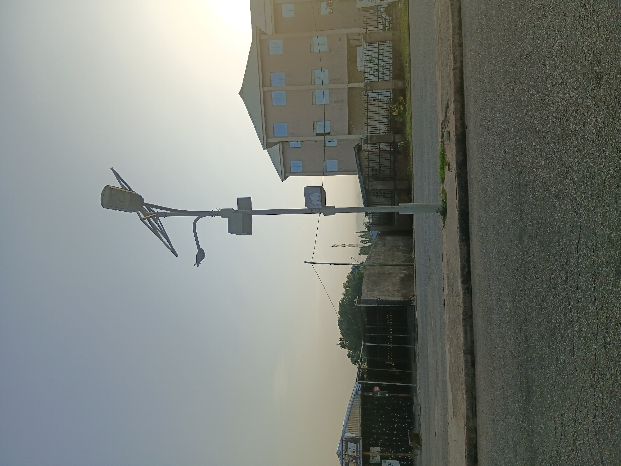 The image depicts a street scene in Jalingo, Nigeria, under a clear sky with a gentle sun low in the frame, suggesting early morning or late afternoon. A prominent street light with a solar panel is positioned in the foreground, to the left of the image center. The street light is a modern design, featuring multiple lamps and a vertical pole with attached signage.
To the left of the street light, a low-lying building with a corrugated metal roof and dark walls, partially obscured by signage, is visible. Further back, a fence encloses an area with more structures. Across the street, on the right side of the image, a multi-story building with a light-colored facade and numerous windows stands prominently. This building appears to be commercial or residential. A black metal fence runs along the front of this property. The street itself is paved with asphalt, showing some signs of wear. Greenery, including trees and grass, is present in the background and along the edges of the properties, suggesting a relatively well-maintained urban environment. The overall scene is calm and unremarkable, capturing a typical street view in the city.