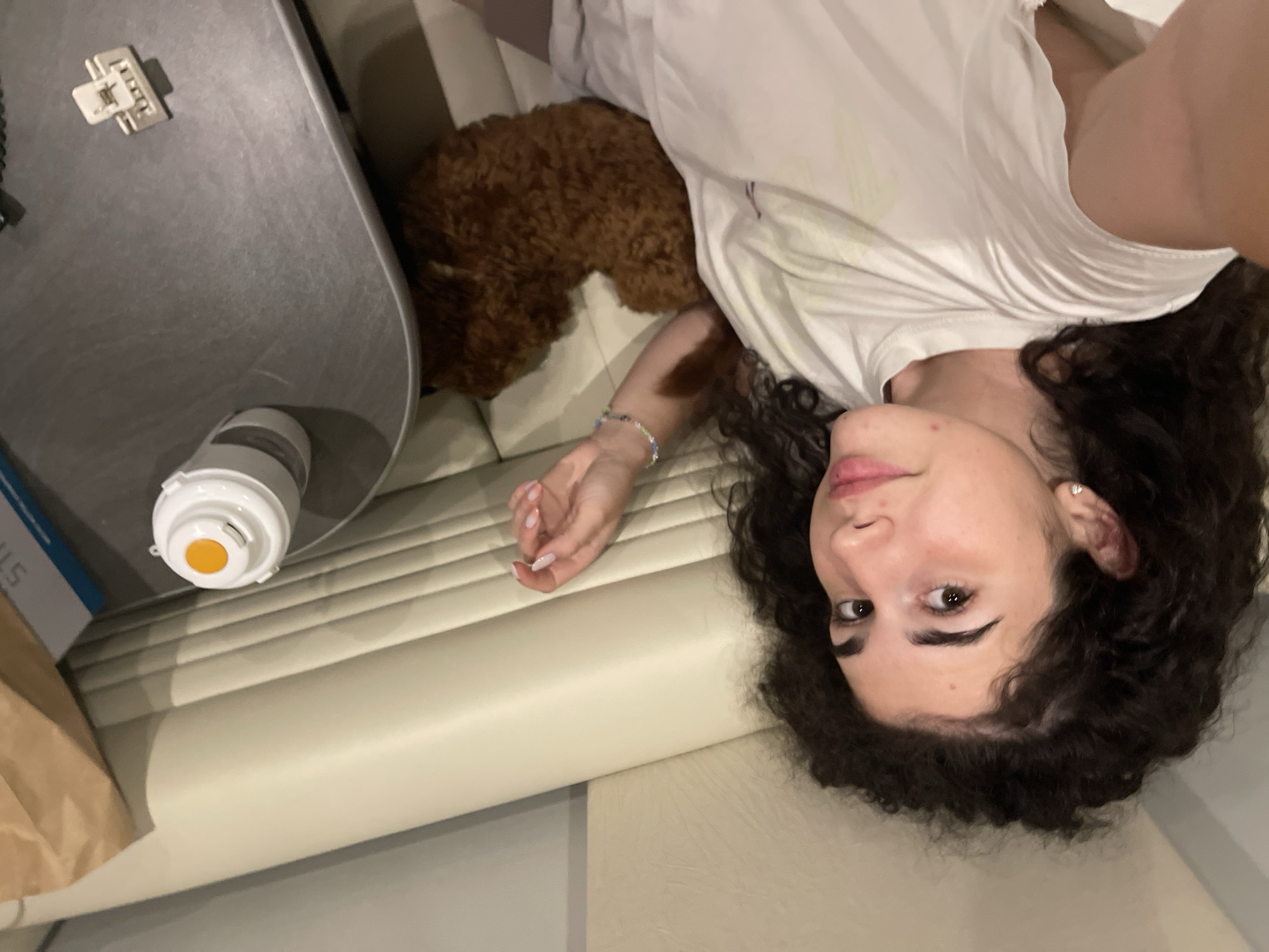 A young female with dark, curly hair and fair complexion is depicted in a close-up, high-angle shot, appearing to be taking a selfie. She is reclining on a light-colored upholstered banquette or booth seating, looking directly towards the camera. She wears a plain white short-sleeved t-shirt and small stud earrings. A multi-colored beaded bracelet is visible on her right wrist, and her fingernails have a light-colored polish. To her right, a dark gray tabletop holds a white insulated cup with an orange cap, a small white hair claw clip, and partially visible objects including a brown paper bag and a blue-edged item (possibly a book or box). A small, brown, curly-haired object, resembling a toy dog or small pet, rests beside her right hip. The seating is beige/cream-colored with horizontal stitching, and a plain light-colored wall or panel is visible behind her. The image provides no discernible visual information to confirm a specific location within the city of Koprinka, Bulgaria; the depicted interior is generic.