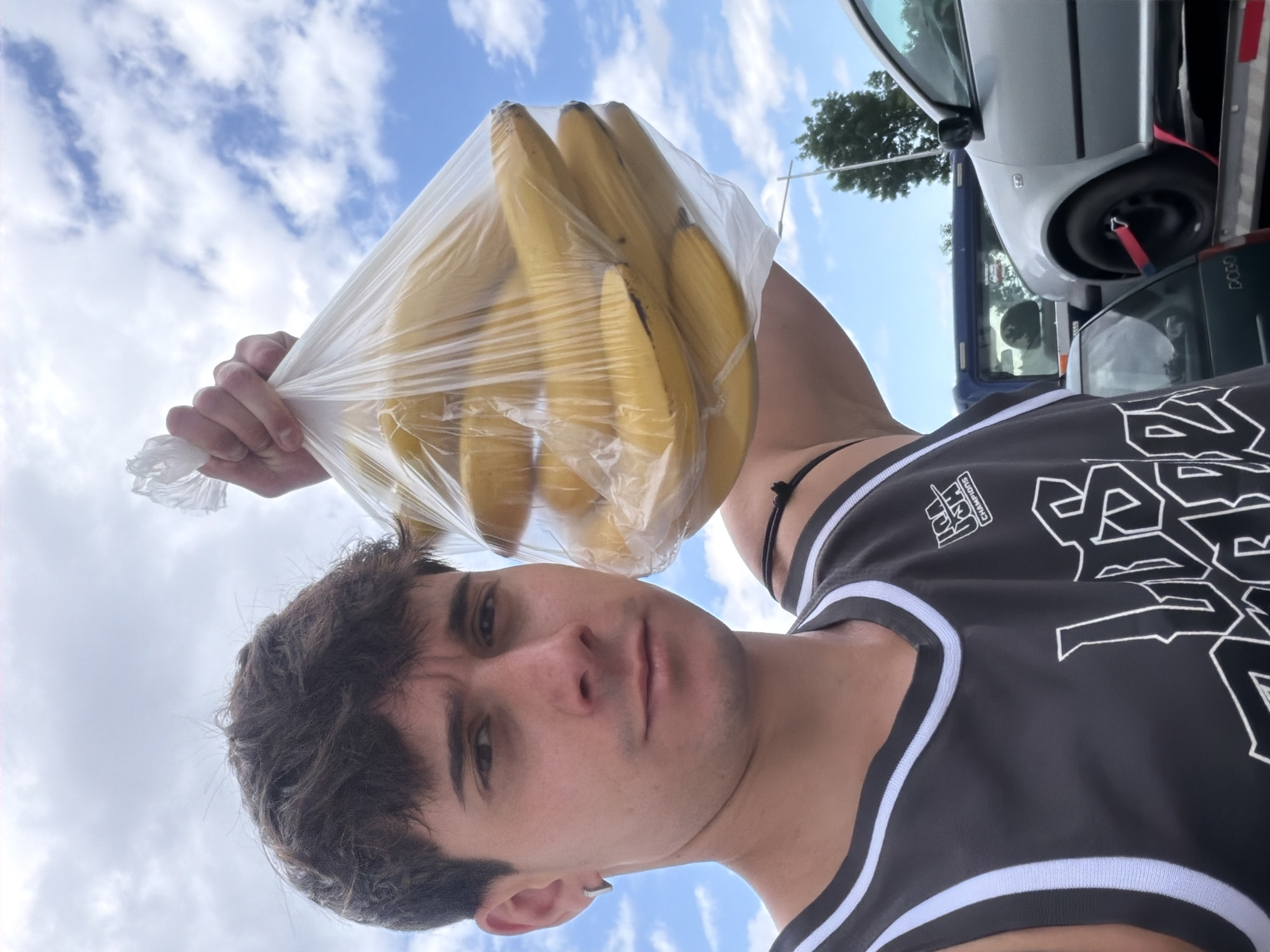 A young man is prominently featured in a low-angle, upright portrait, captured outdoors under a bright, partly cloudy blue sky, likely in Sofia, Bulgaria. He has dark hair, light stubble, and a neutral to slightly serious expression, looking directly into the camera. He wears a black tank top or basketball jersey with white trim along the neckline and armholes. On his left shoulder, white text reads "J&M CHAMPIONS," and a larger, stylized white graphic is visible on the front of the jersey. He also wears a small silver hoop earring in his left ear and a black cord necklace.
With his right hand, the man holds a transparent plastic bag containing a bunch of yellow bananas, elevated against the sky. To the right side of the frame, parts of two vehicles are visible: a silver car, possibly a hatchback, with a red strap near its tire, sits above a darker vehicle. The word "DOJO" is discernible on the side of the darker vehicle. A tree is also partially visible in the upper right background. The image appears to be a daytime shot, indicating pleasant weather, possibly taken as a selfie or a close-up photo in an urban or suburban setting.