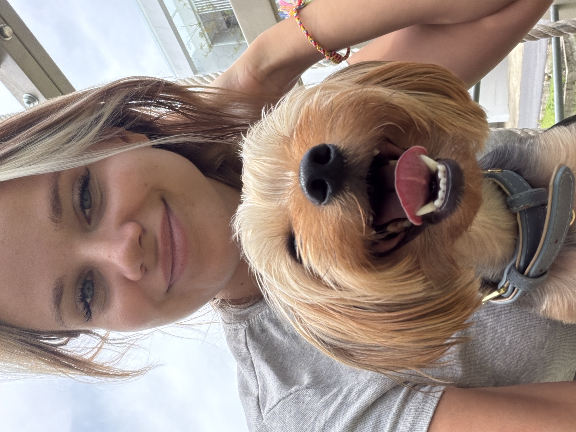 A young woman with blonde hair, styled with highlights, is taking a selfie with a small, tan-colored dog. The dog is held in front of the woman, with its face prominently displayed and mouth open in a happy expression, revealing its tongue and teeth. The woman is wearing a grey t-shirt. Her left arm is visible, resting on the dog. A colorful woven bracelet is visible on her right wrist. The background appears to be outdoors, with a cloudy sky and a hint of modern architecture, possibly a building with balconies, suggesting an urban or suburban environment. The image seems to have been taken during daylight. The location is identified as Thep Krasatti, Thailand.