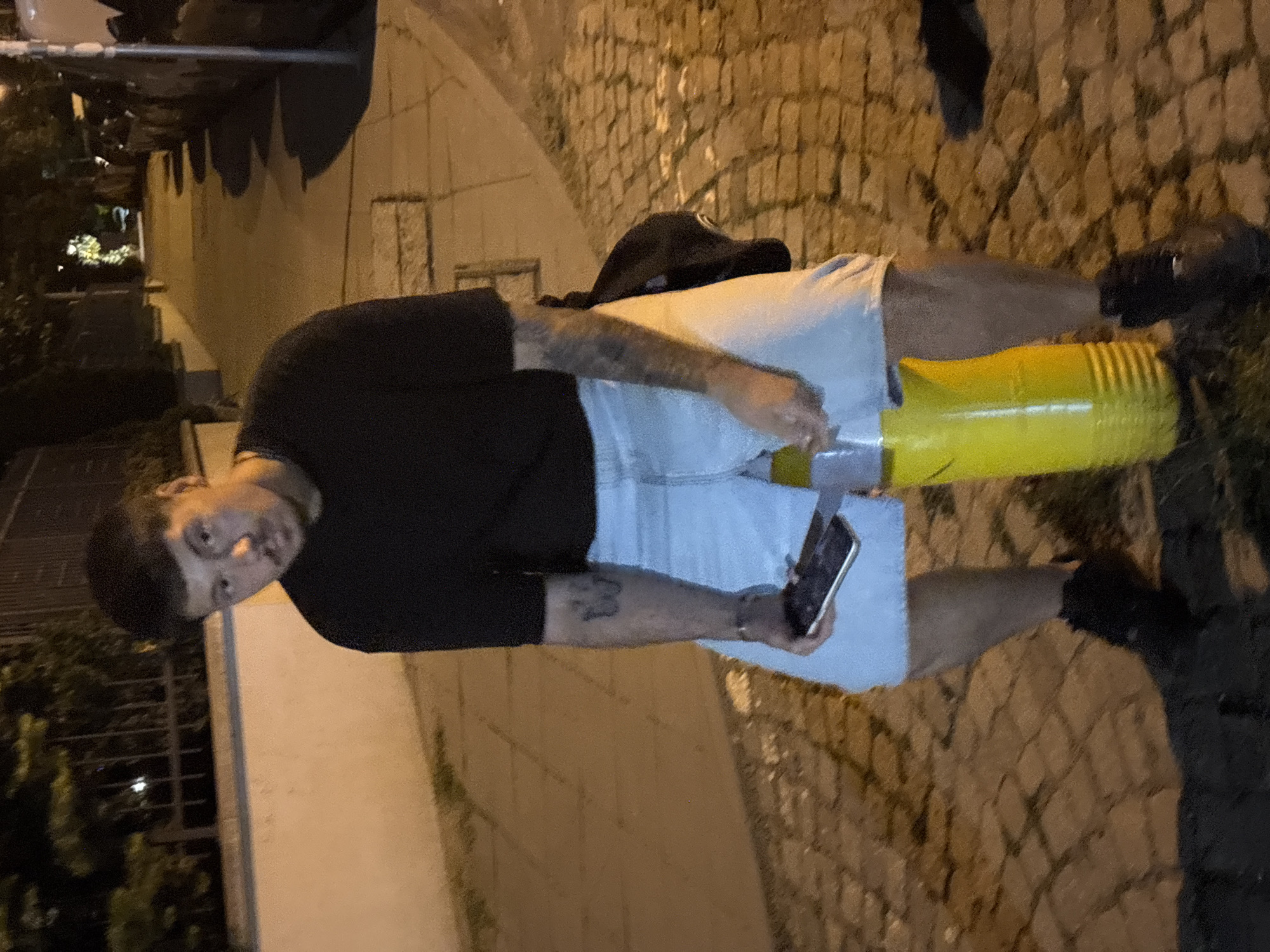 A man is standing on a cobblestone street in Sofia, Bulgaria. He is holding a smartphone in his left hand and a yellow object in his right hand. He is wearing a black shirt and light-colored shorts. A black baseball cap and a black bag are resting on the yellow object behind him. The background includes street lamps and buildings.