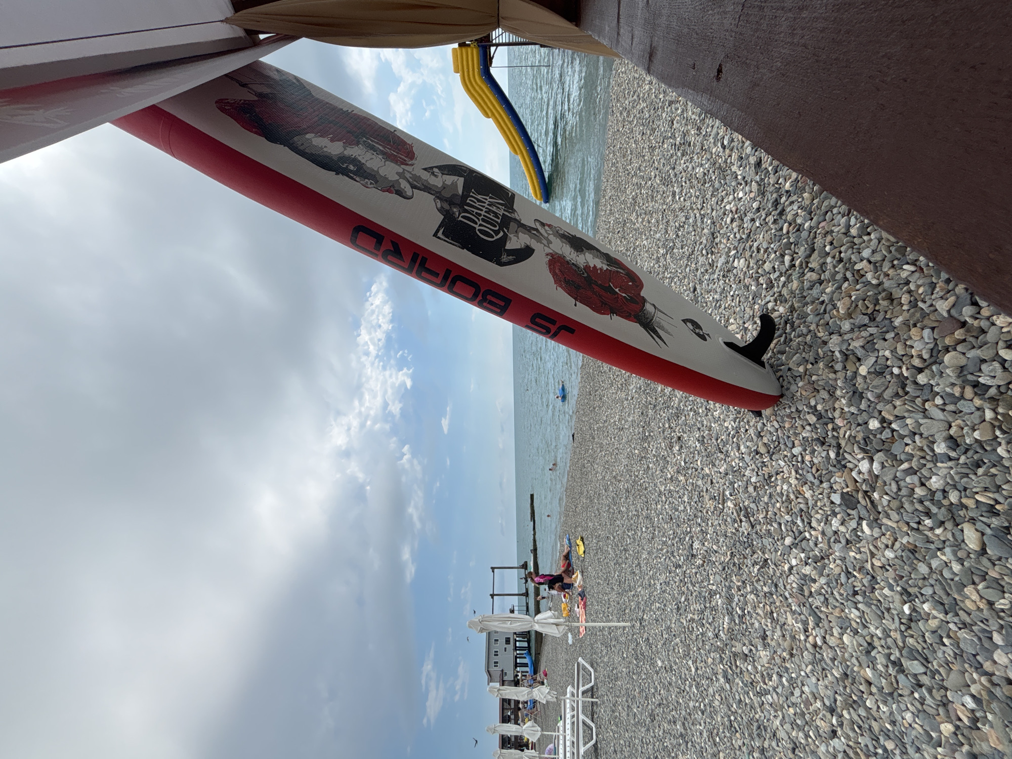 This media file depicts a beach scene in Sochi, Russia, with a focus on a stand-up paddleboard in the foreground. The paddleboard, labeled "JS BOARD" and featuring the graphic "DARK QUEEN," is white with a red stripe and a black fin. It is positioned diagonally, partially obscured by a tan canopy overhead and a dark wooden beam.
The background reveals a pebbled beach leading to the Black Sea. Several beach umbrellas and lounge chairs are visible, along with a few people sunbathing and one person in the water. A yellow and blue water slide is partially visible near the sea. The sky is partly cloudy, suggesting it is daytime. The overall impression is a relaxed summer day at the beach, with opportunities for water activities.