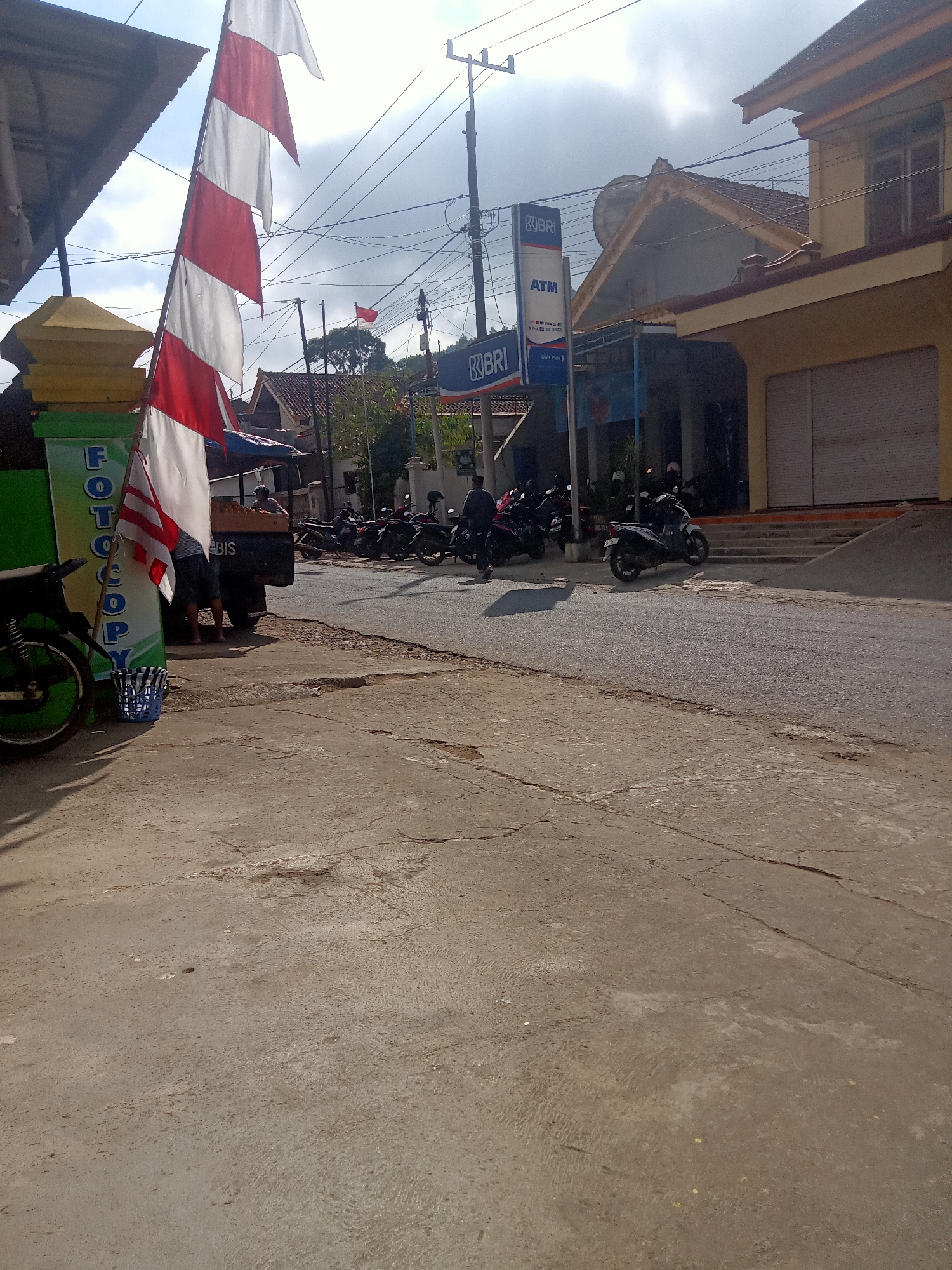 The image depicts a street scene in Pule, Indonesia, featuring a prominent red and white Indonesian flag fluttering from a pole. The flag is positioned to the left of the frame, with several horizontal red and white stripes. In the background, there is a building with a yellow facade and a closed shuttered entrance. A blue and white ATM sign for BRI is visible on a pole in front of the building. Several motorcycles are parked along the roadside, and some individuals are standing near a truck with a canopy. The ground is paved with concrete, showing some cracks and wear. The sky is cloudy with patches of sunlight. To the left of the flag, there's a green sign with the word "FOTOCOPY" printed vertically in white lettering. A blue woven basket sits on the ground near the flag.