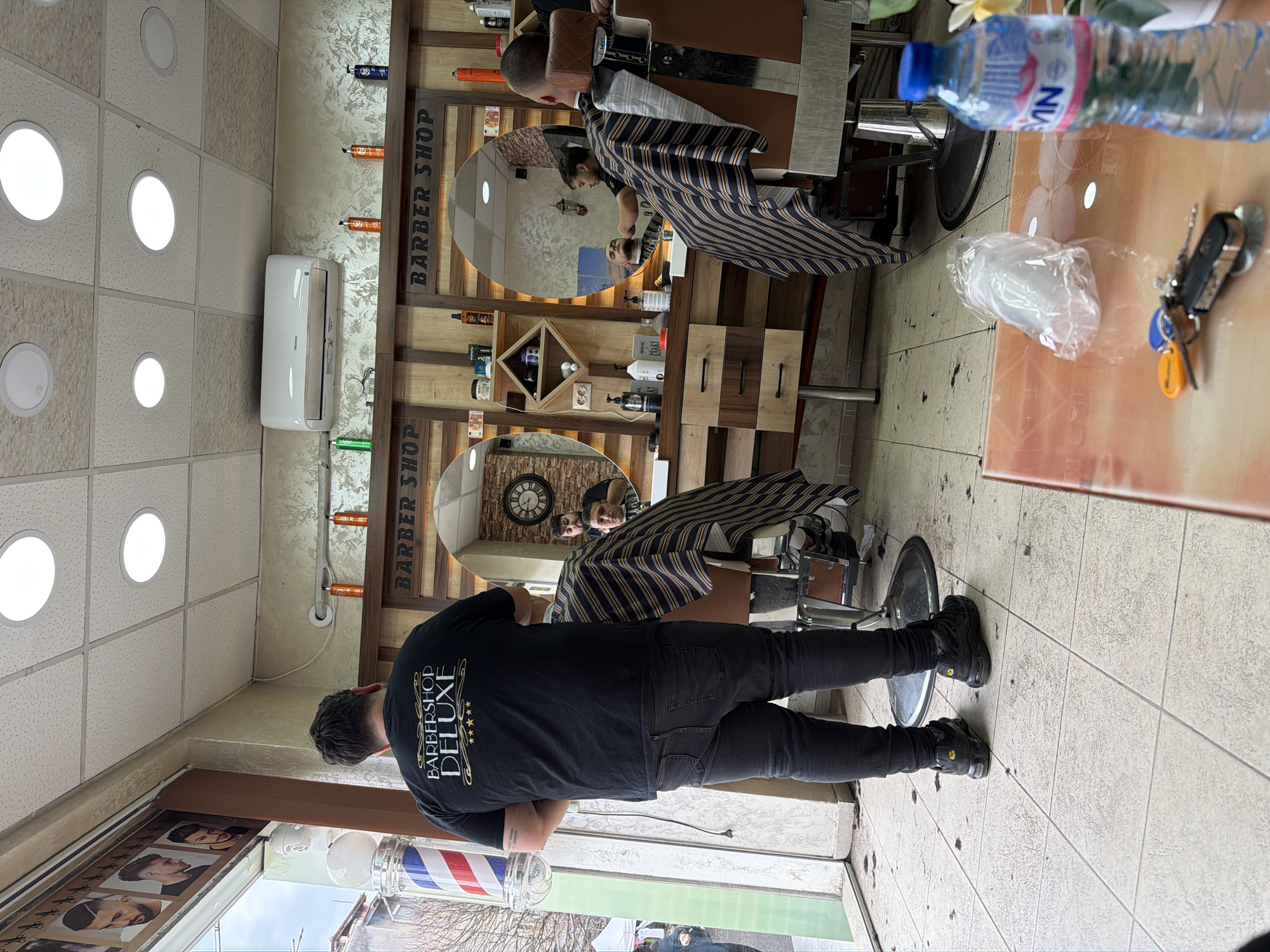 The image depicts the interior of a barbershop, presumably in Panagyurishte, Bulgaria. A man in a black t-shirt with "BARBERSHOP DELUXE" printed on the back stands near the entrance, facing towards two barber chairs occupied by customers receiving haircuts. The barbershop is decorated with a wood-paneled wall featuring the words "BARBER SHOP" and a clock. The floor is tiled, and there are cut hairs on the floor around the chairs. A table to the right holds a bottle of water, a plastic bag, and keys. The entrance is visible through the window, and a traditional barber pole is also visible.