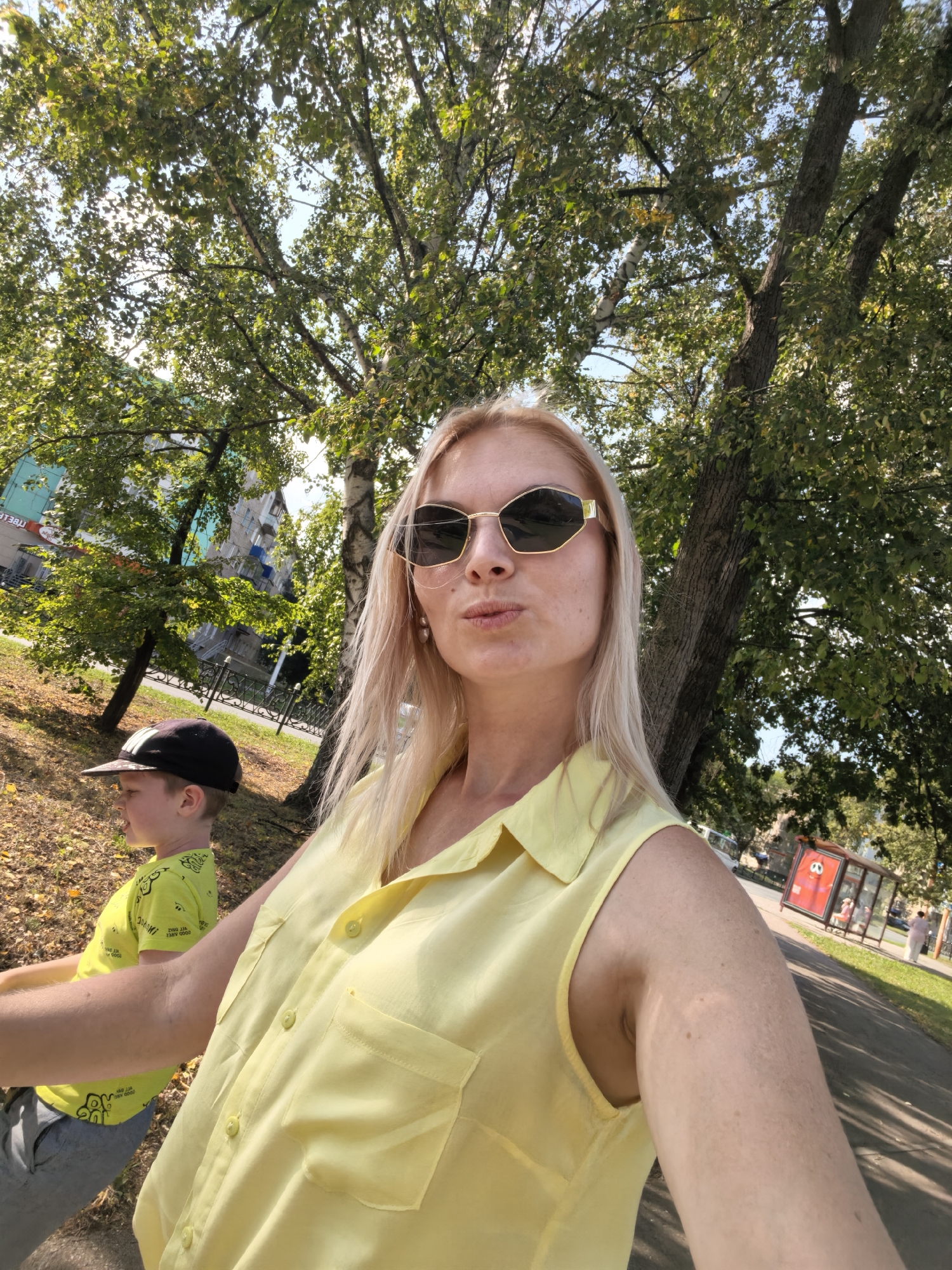 In Sterlitamak, Russia, on a sunny day, a woman with blonde hair and sunglasses is taking a selfie outdoors. She is wearing a yellow sleeveless shirt. To her left, a young boy wearing a black cap and a neon yellow t-shirt is visible. They are in a park-like setting with trees and a paved pathway. In the background, there are buildings and a bus stop shelter.