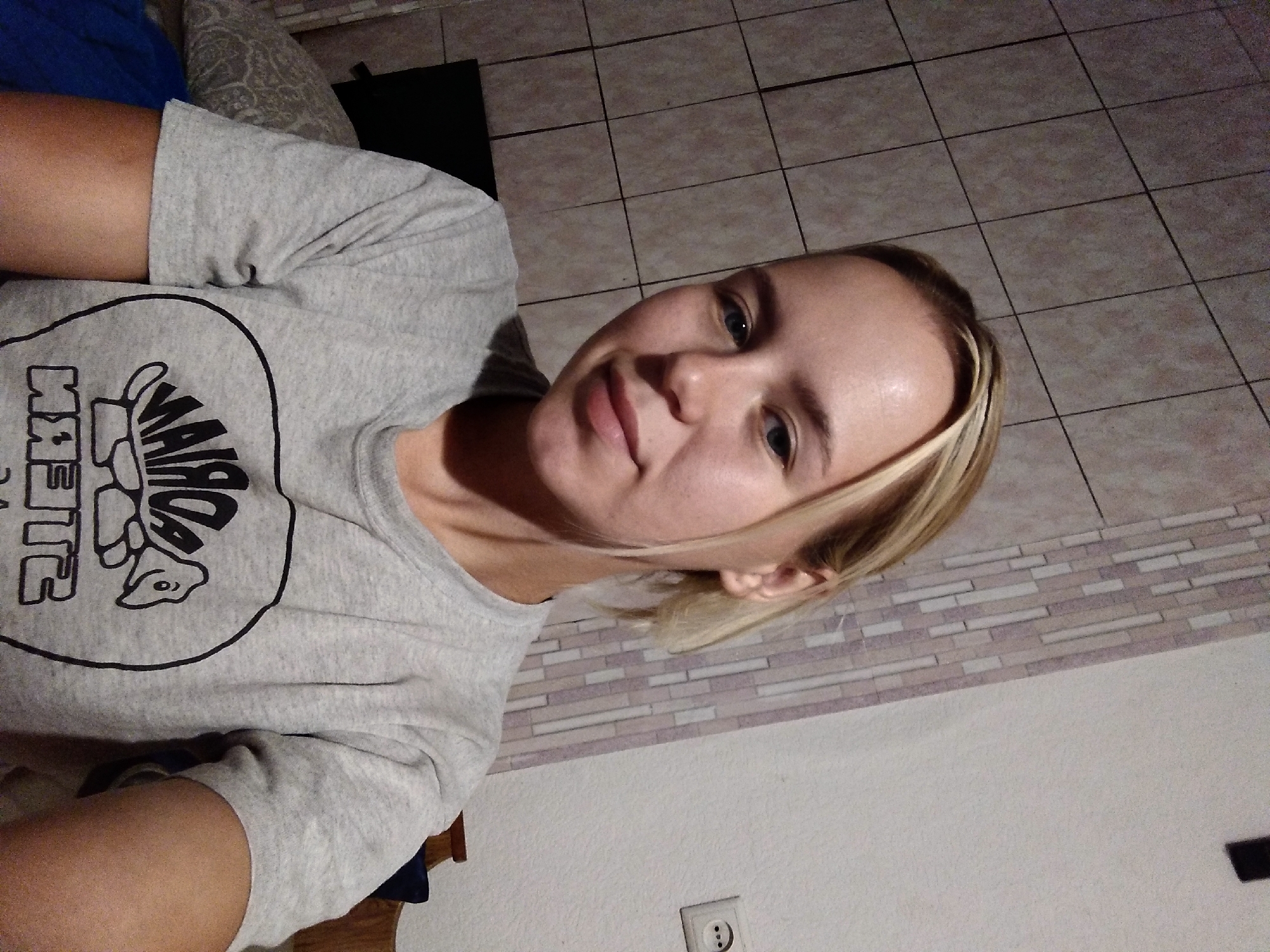 A young Caucasian woman with blonde hair pulled back is shown in a medium shot, looking directly at the camera. She is wearing a grey t-shirt with a black circular graphic on the front. The background consists of a tiled wall and a plain wall with an electrical outlet. The scene appears to be indoors, likely in a residential setting.