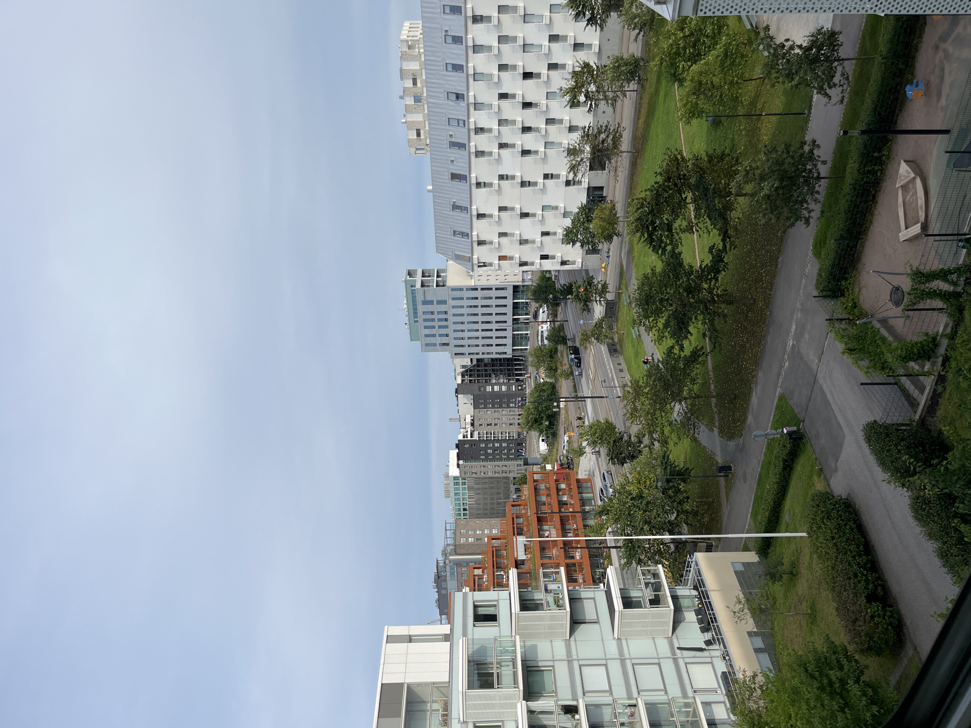 The image shows a view of a modern residential area in Helsinki, Finland, on an overcast day. In the foreground, a light blue apartment building with glass balconies dominates the left side. To its right, a building with orange brick facades features multiple balconies. Further back, several taller apartment buildings of varying architectural styles are visible. One prominent white building with a metal roof has a grid of many evenly spaced windows. Another building is characterized by its tall, narrow structure with numerous windows. Beyond these, a cityscape with denser buildings and structures extends into the distance.
In the middle ground, a street runs horizontally with cars and pedestrians. A green park area with scattered trees and pathways is situated between the street and the apartment buildings. In the foreground, to the lower right, a playground with a sandbox and swing is visible. The environment appears to be a well-maintained urban area with a mix of residential buildings and green spaces.